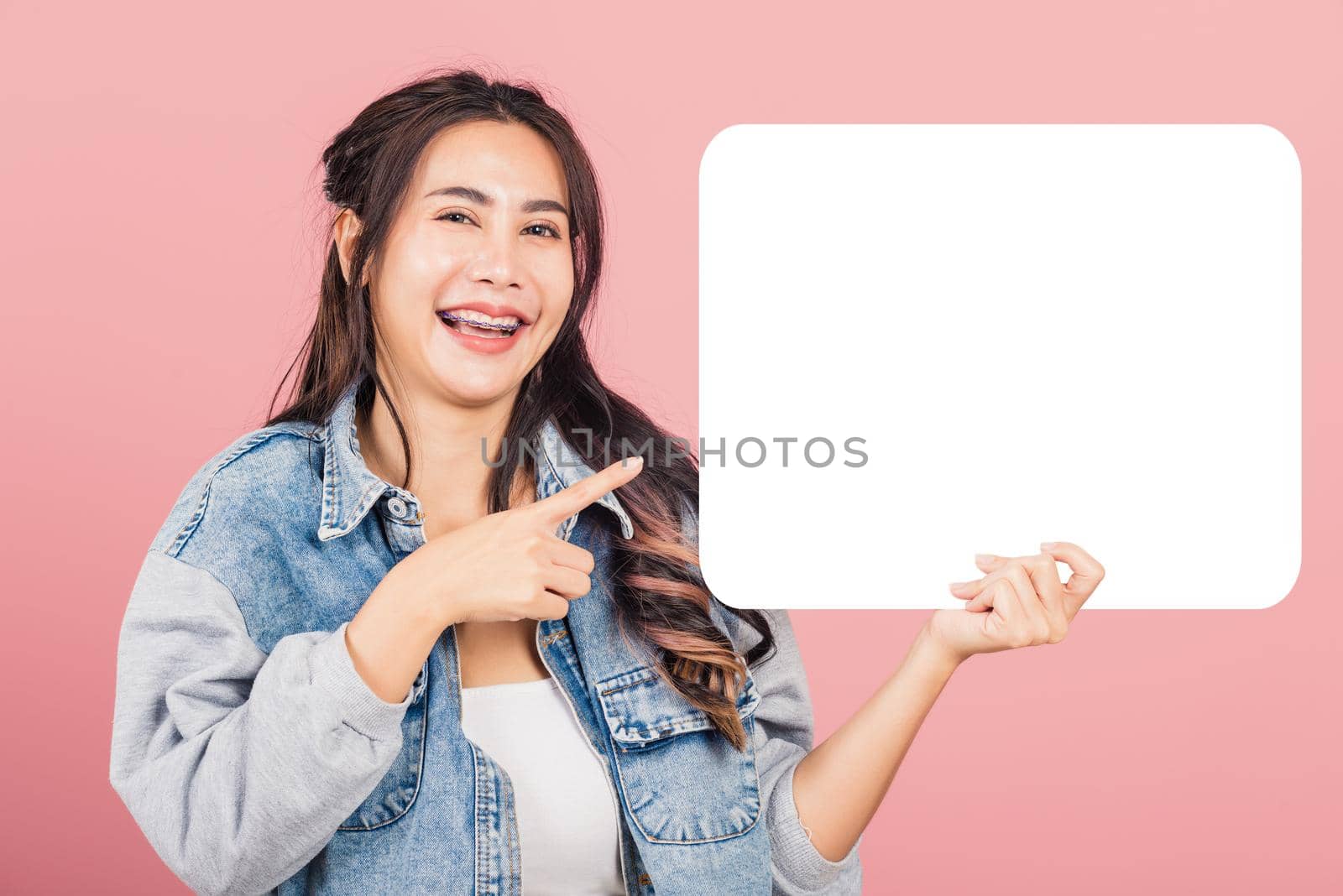 Happy Asian beautiful young woman smiling excited wear denims hold empty speech bubble sign, Portrait female posing finger pointing bubble for your text idea, studio shot isolated on pink background