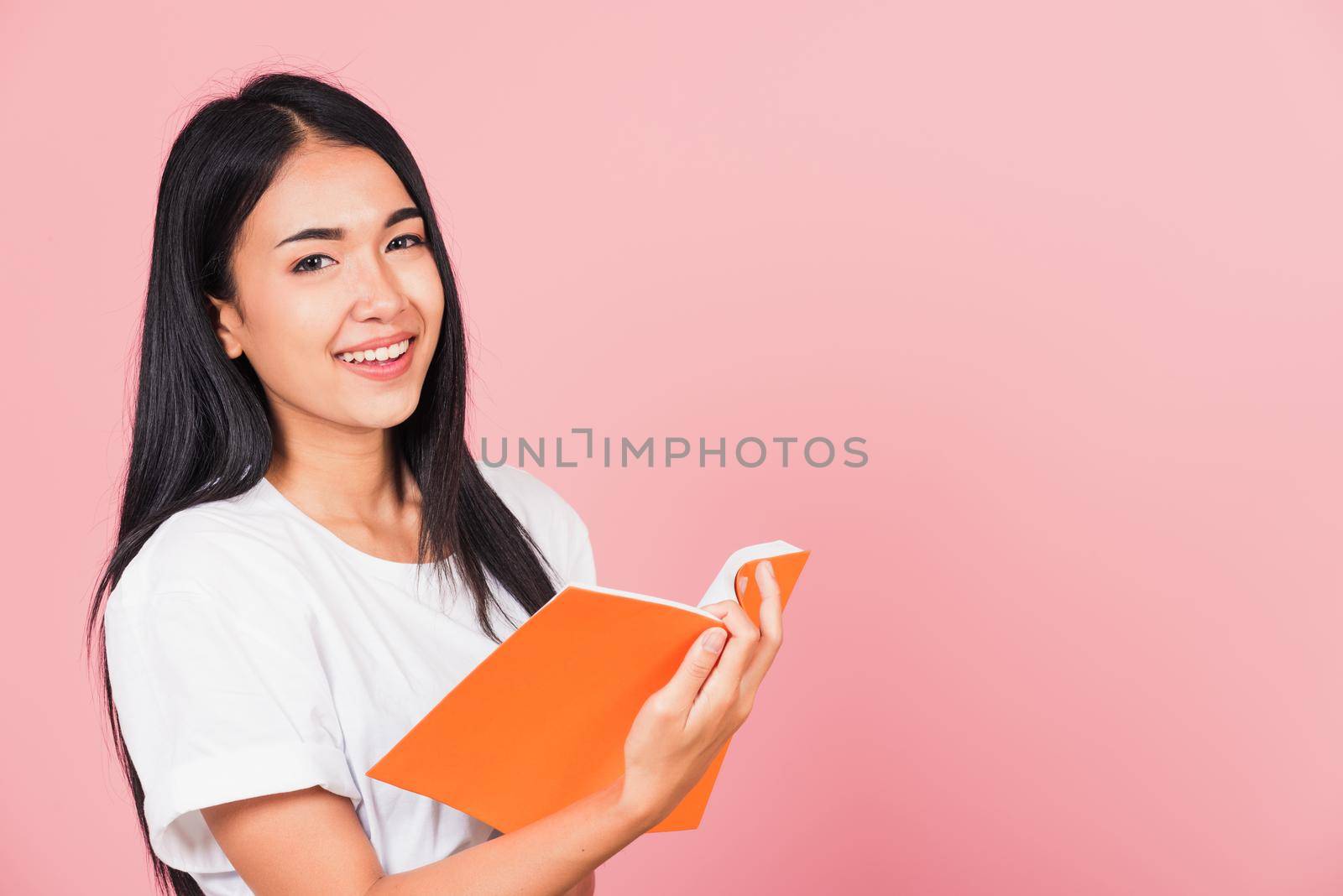 Portrait of happy Asian beautiful young woman confident smiling standing holding orange book open or diary for reading, studio shot isolated on pink background, with copy space, education concept