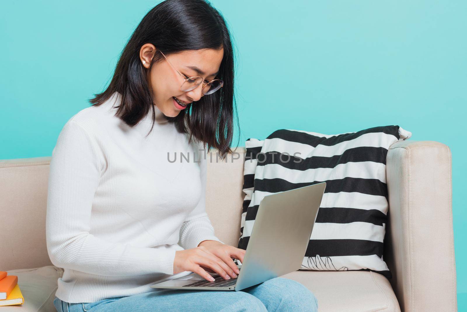 Asian beautiful young woman wear eyeglasses sitting on sofa using a laptop, portrait relaxation of happy female smiling in living freelancer working on computer studio shot isolated on blue background