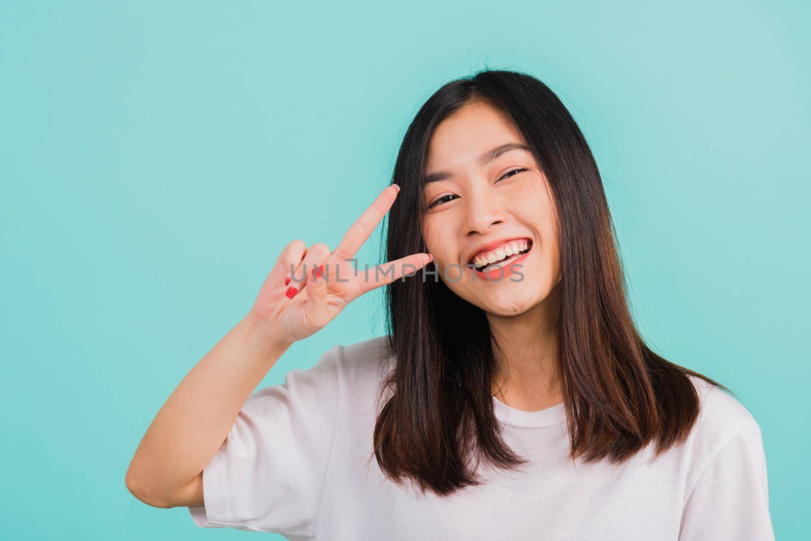 Asian happy portrait beautiful cute young woman teen smile standing wear t-shirt showing finger making v-sign symbol near eye looking to camera isolated, studio shot blue background with copy space