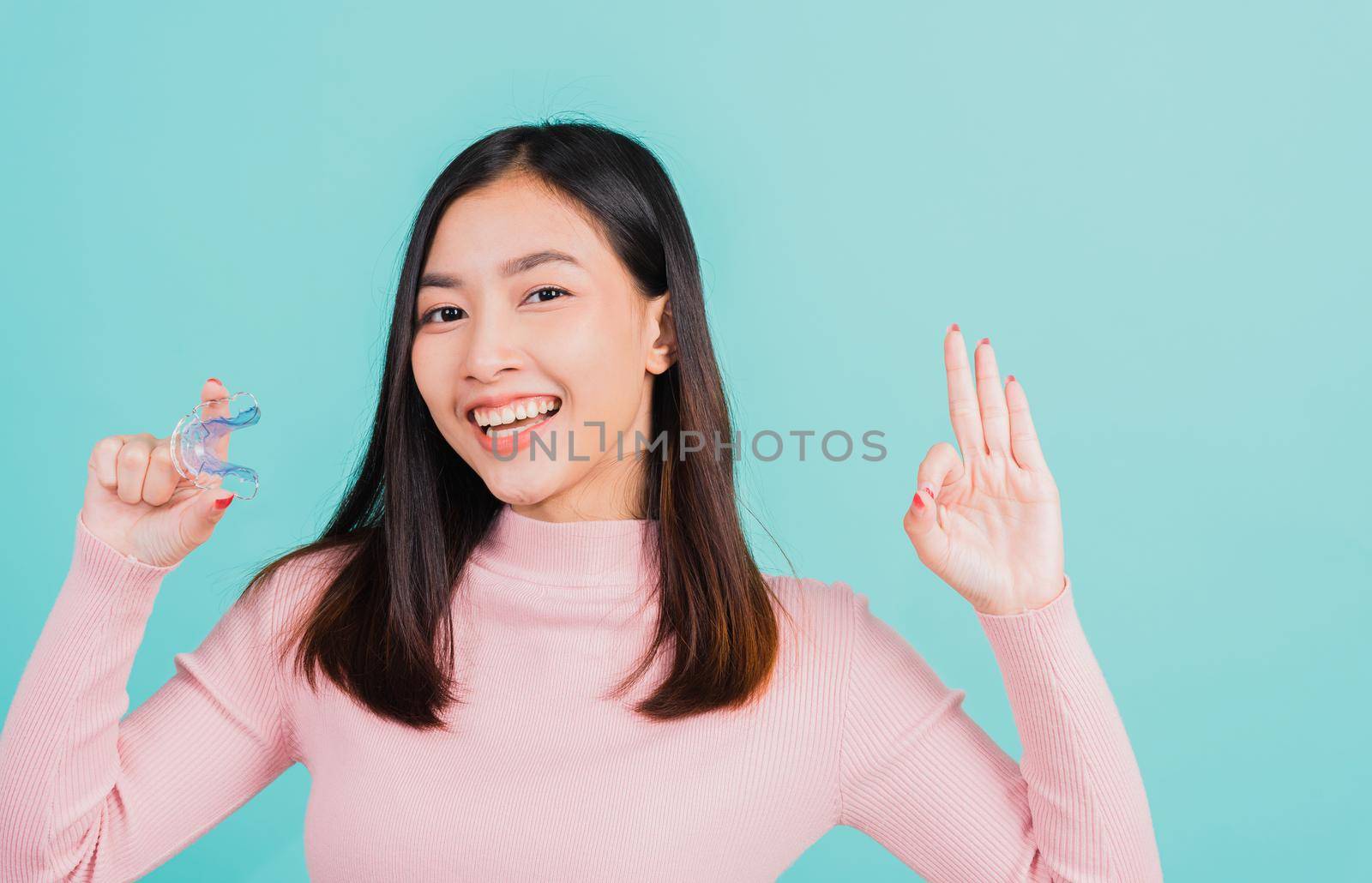 Portrait Asian beautiful young woman smiling holding silicone orthodontic retainers for teeth and show finger brackets saying OK sign, isolated on blue background, Dental hygiene healthy care concept