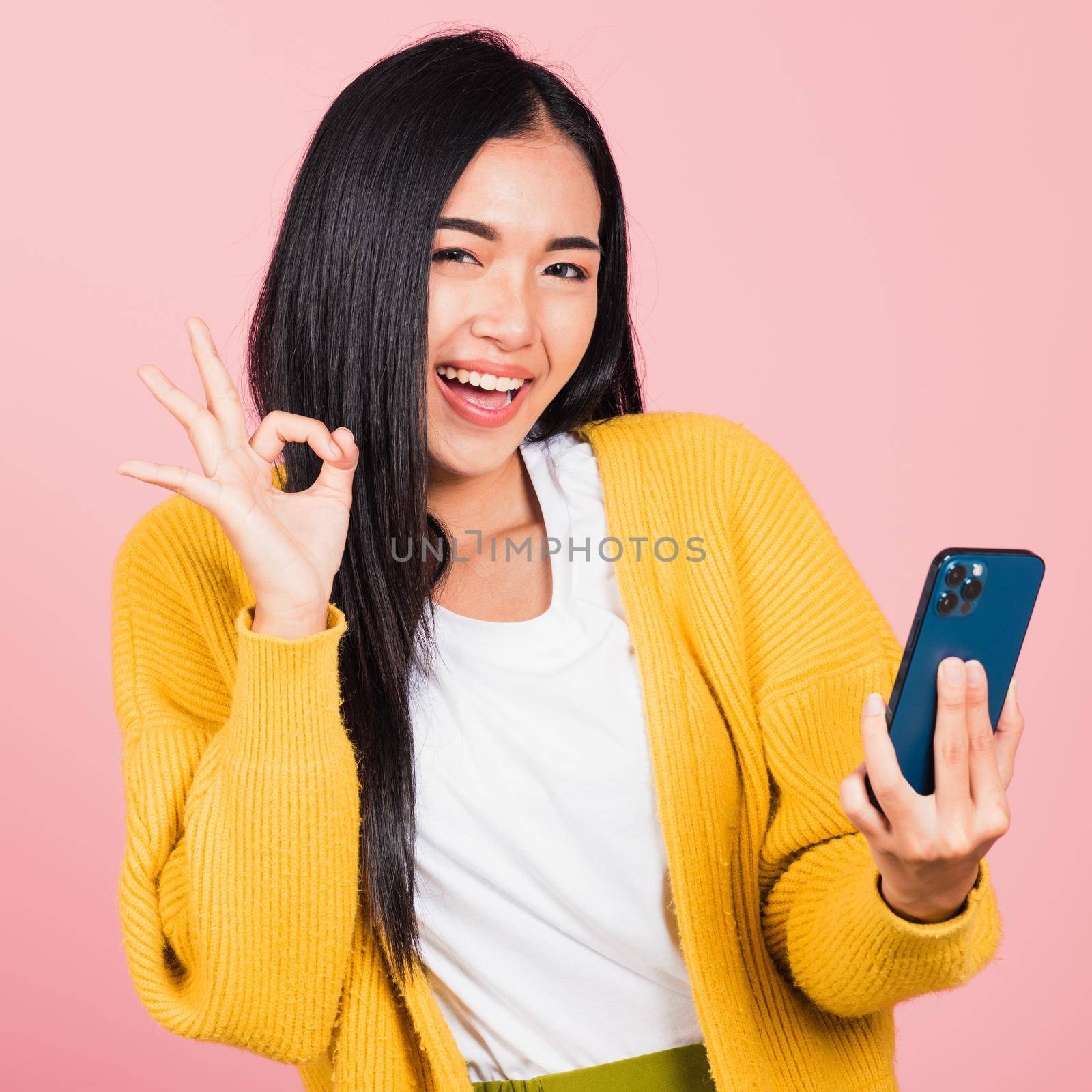 Happy Asian portrait beautiful cute young woman excited holding mobile phone and gesturing ok sign, studio shot isolated on pink background, Thai female making finger symbol on smartphone