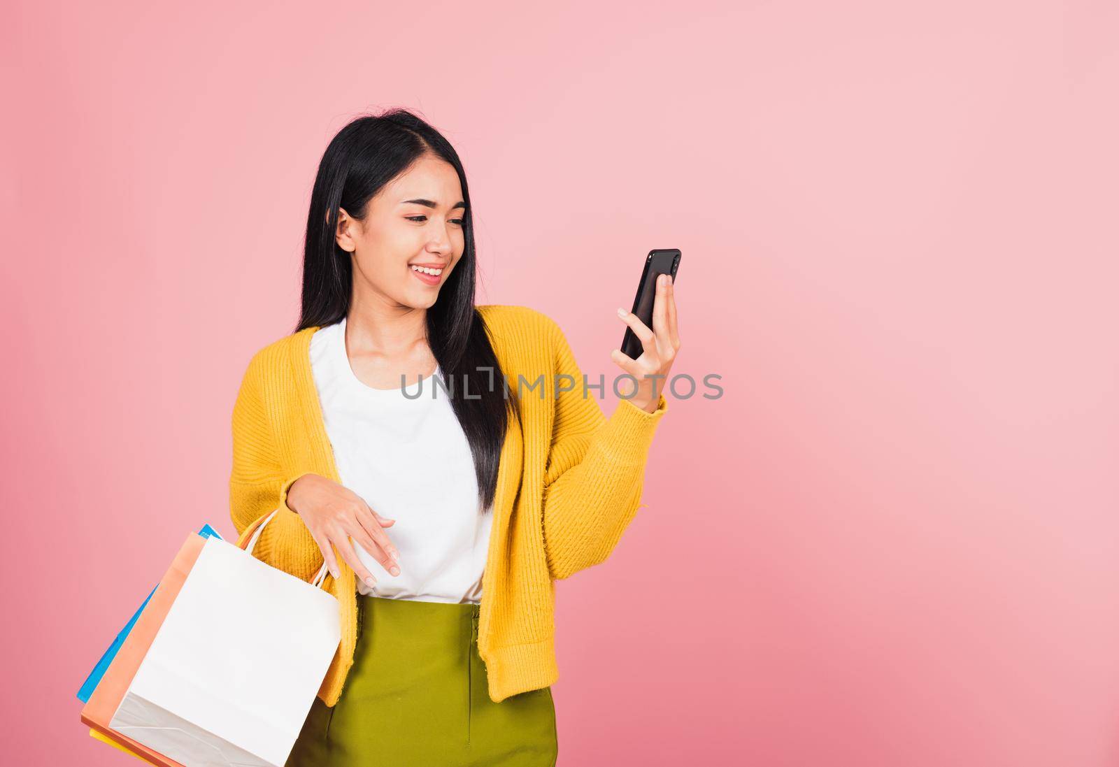 Portrait Asian happy beautiful young woman teen shopper smiling standing excited holding online shopping bags online colorful multicolor and smartphone on hand, studio shot isolated on pink background