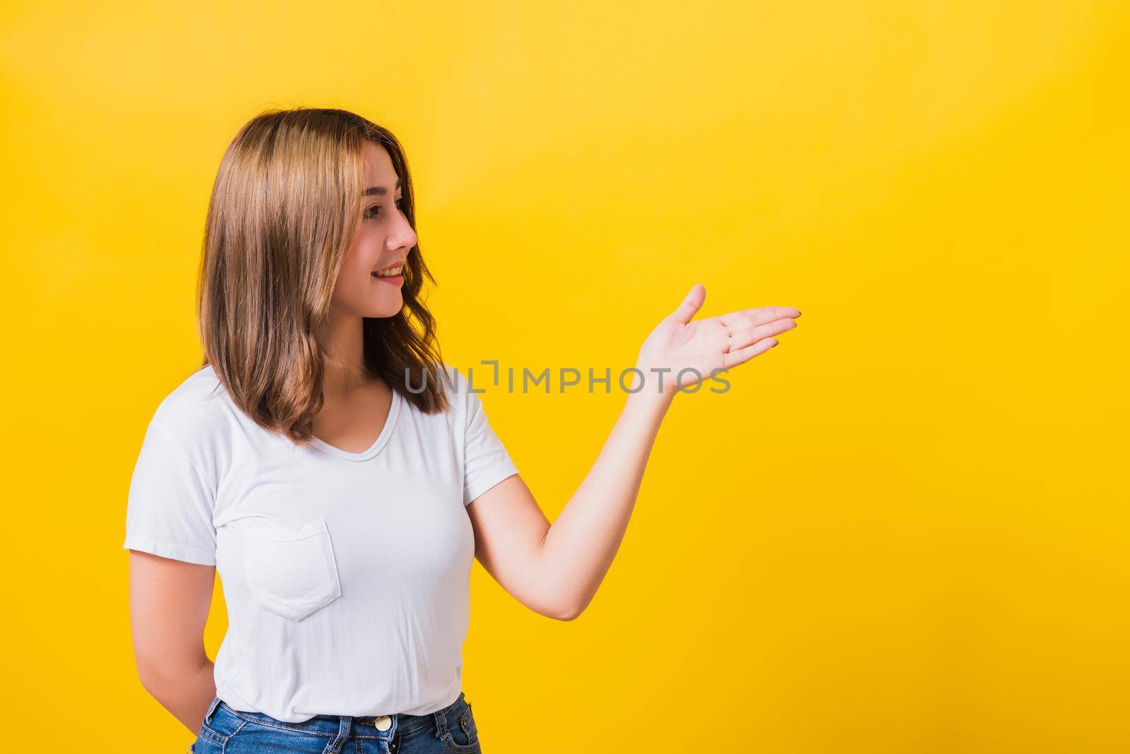 Asian Thai happy portrait beautiful cute young woman standing wear white t-shirt holding something on palm away side looking to side, studio shot isolated on yellow background with copy space