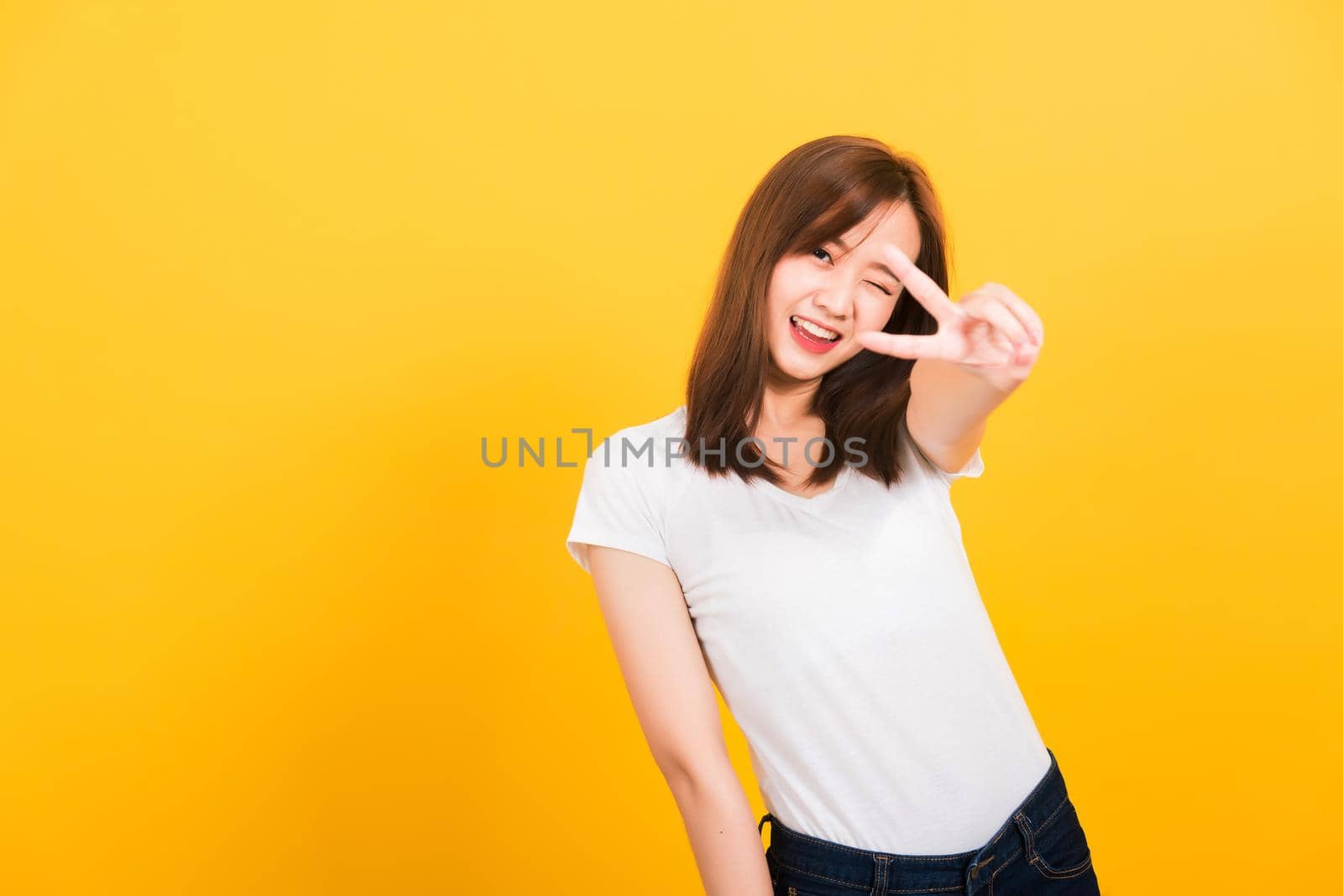 Asian happy portrait beautiful cute young woman teen smile standing wear t-shirt showing finger making v-sign symbol near eye  looking to camera isolated, studio shot yellow background with copy space