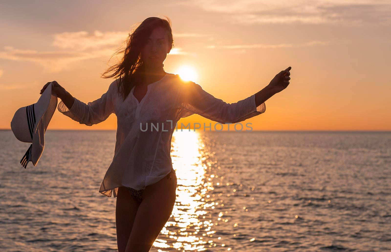 Silhouette of young happy carefree woman posing with hat on the beach at sunset
