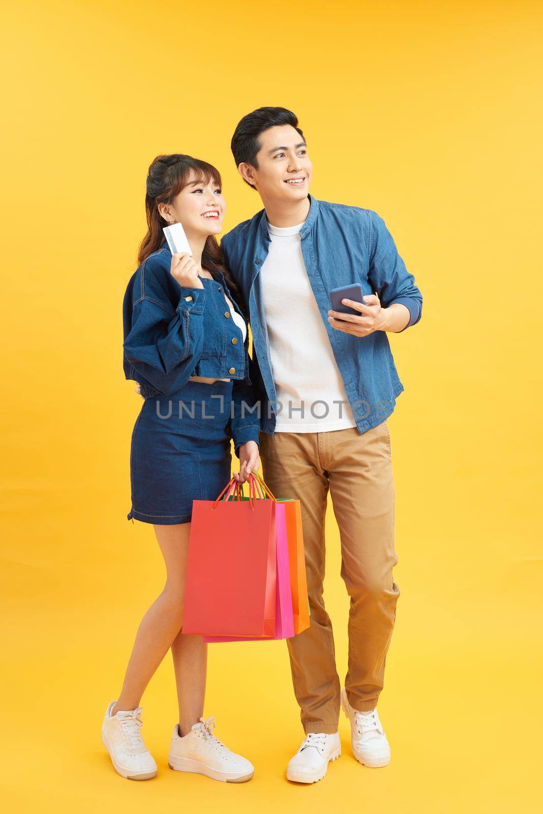 portrait of smiling couple holding shopping bags and credit card isolated on yellow by makidotvn