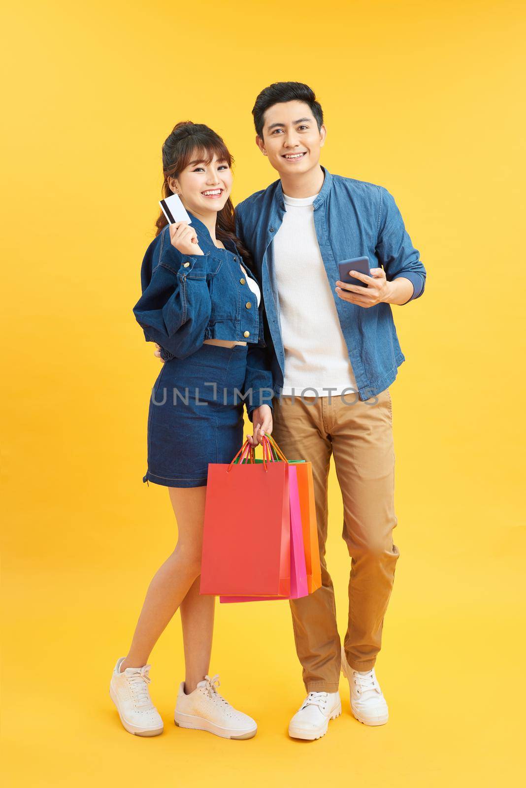portrait of couple holding shopping bags and credit card isolated on yellow by makidotvn
