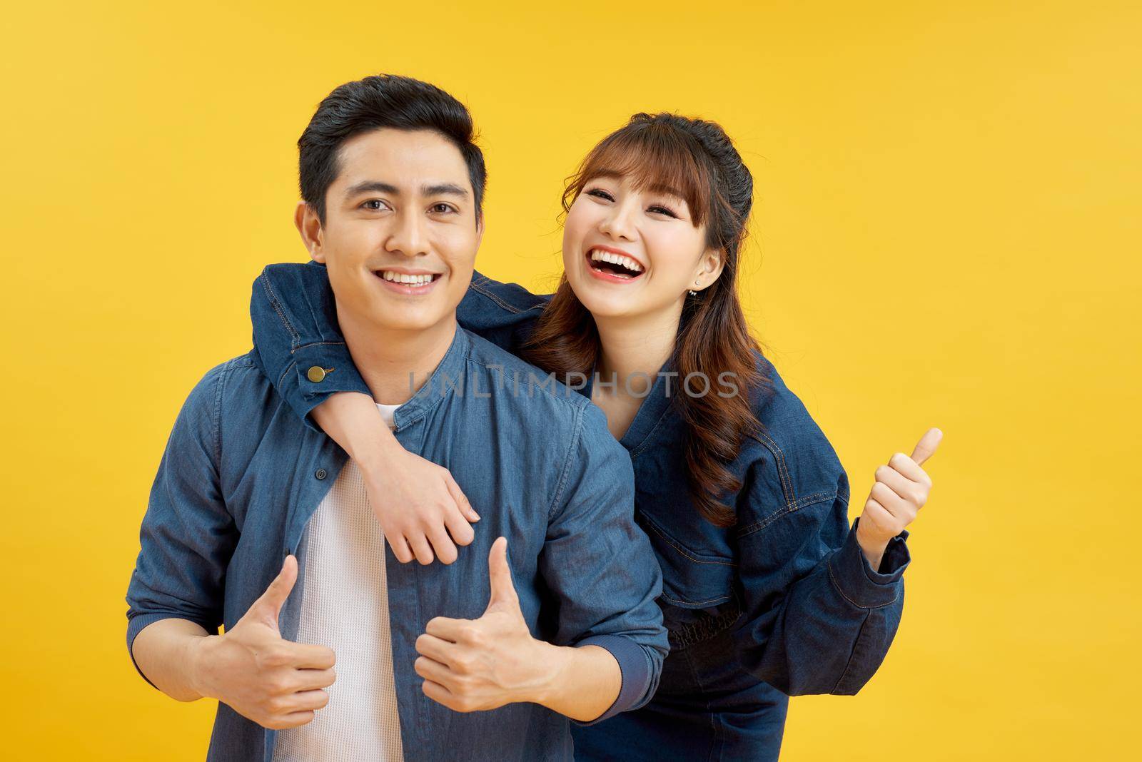 Portrait of cheerful people man and woman in basic clothing smiling and showing thumb up at camera isolated over yellow background