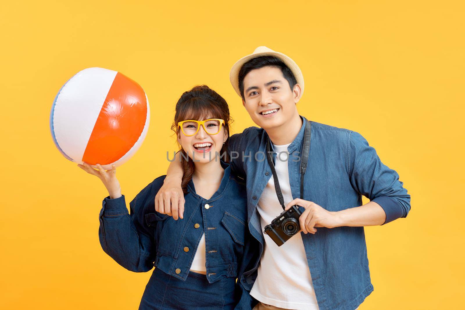 Young smiling happy couple tourists in casual summer attire with beach accessories on colorful yellow studio background by makidotvn