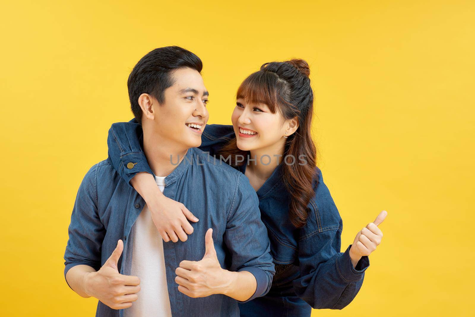 Portrait of smiling couple man and woman showing thumb up together while doing piggyback ride isolated over yellow background