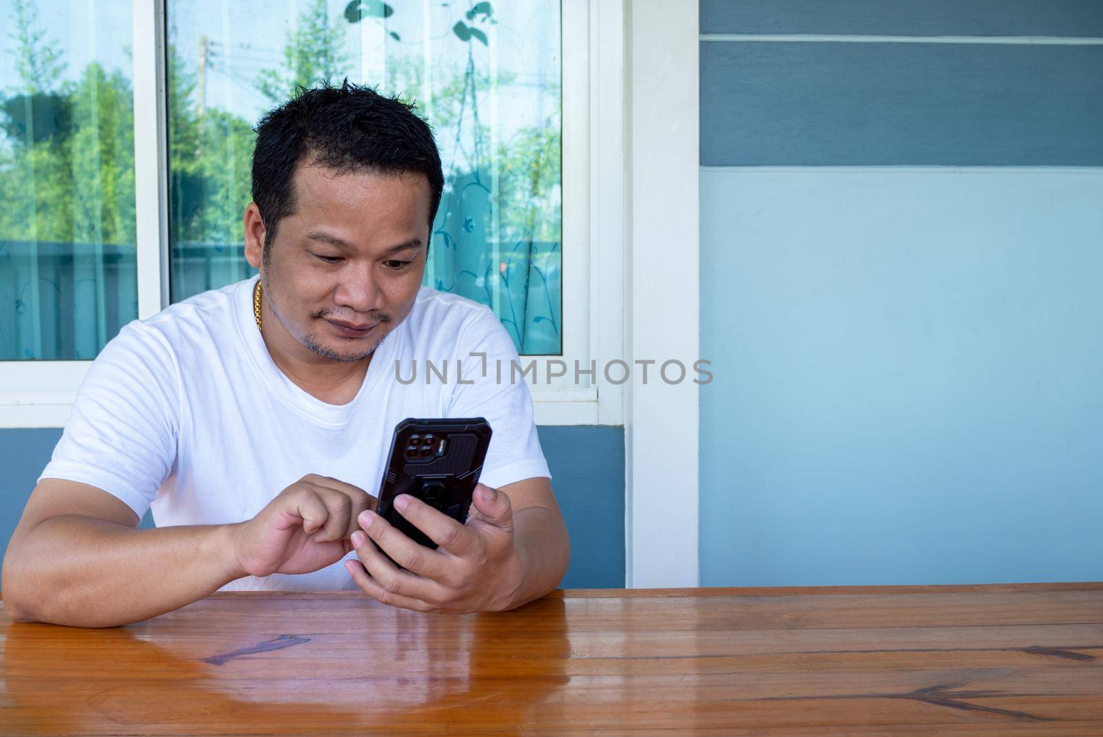 Asian man wearing white shirt using the phone on a wooden table by Unimages2527