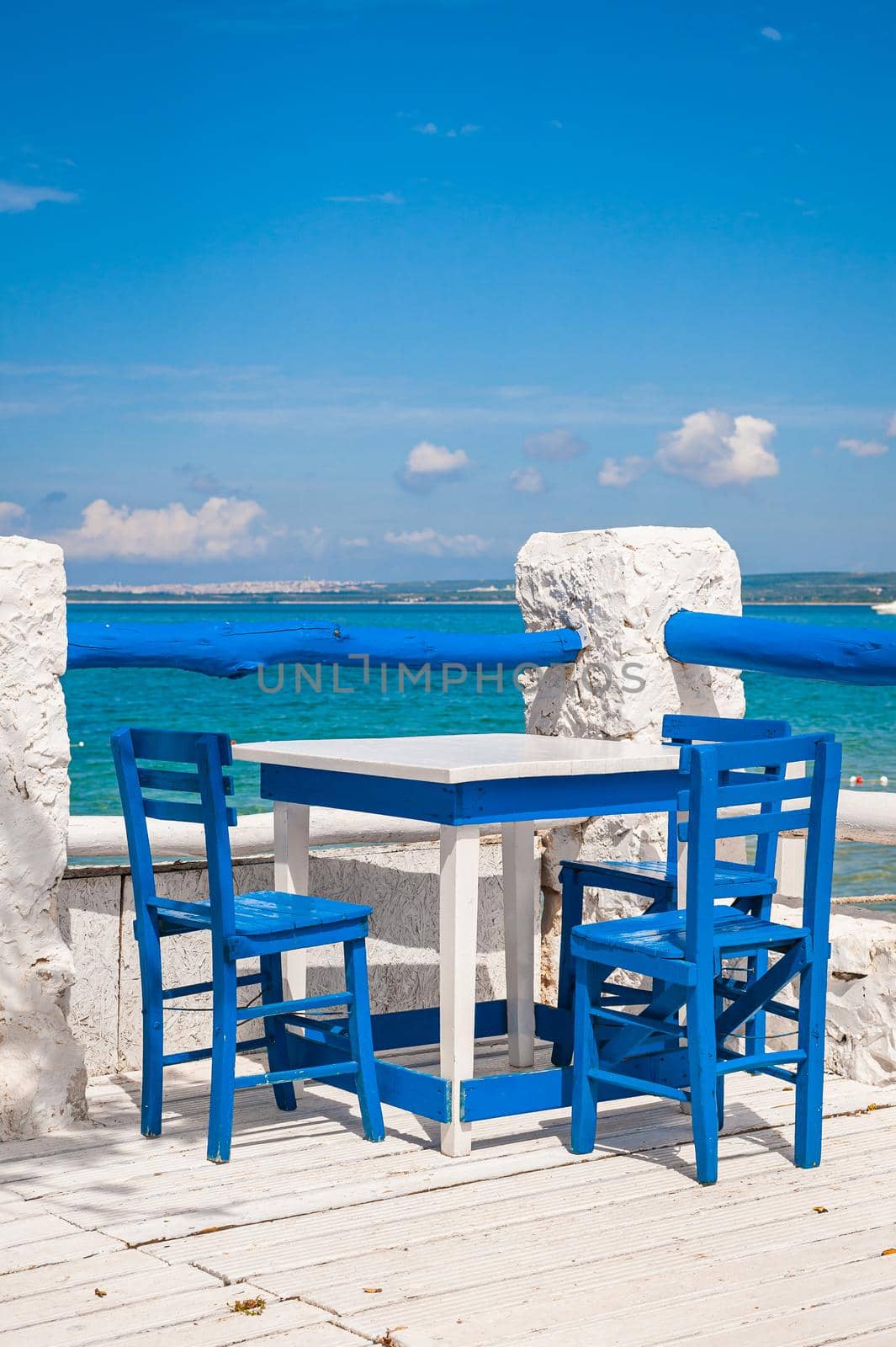 Wooden blue chairs and white tables on seaside terrace with a view on calm waters.