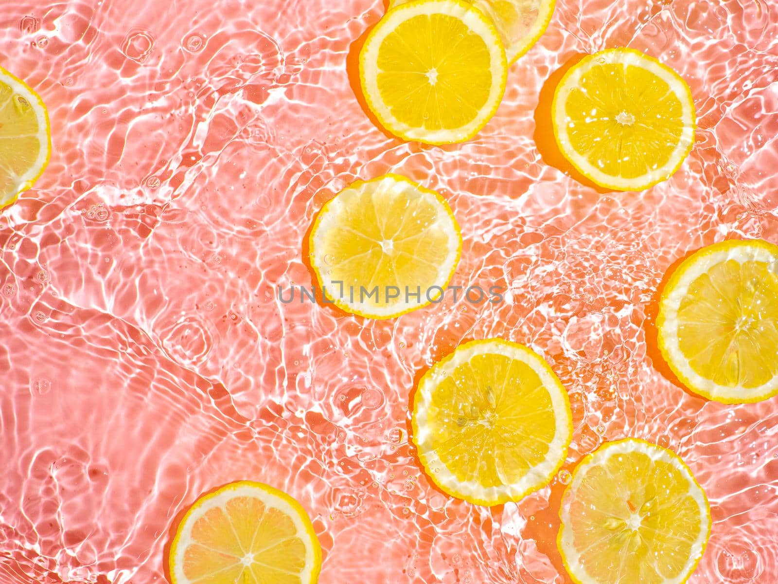 Lemon slices in clean transparent water over pink background with copy space. Water splashing on water surface in sunlight. Top view or flatlay. Summer, vacation, healthy eating concept