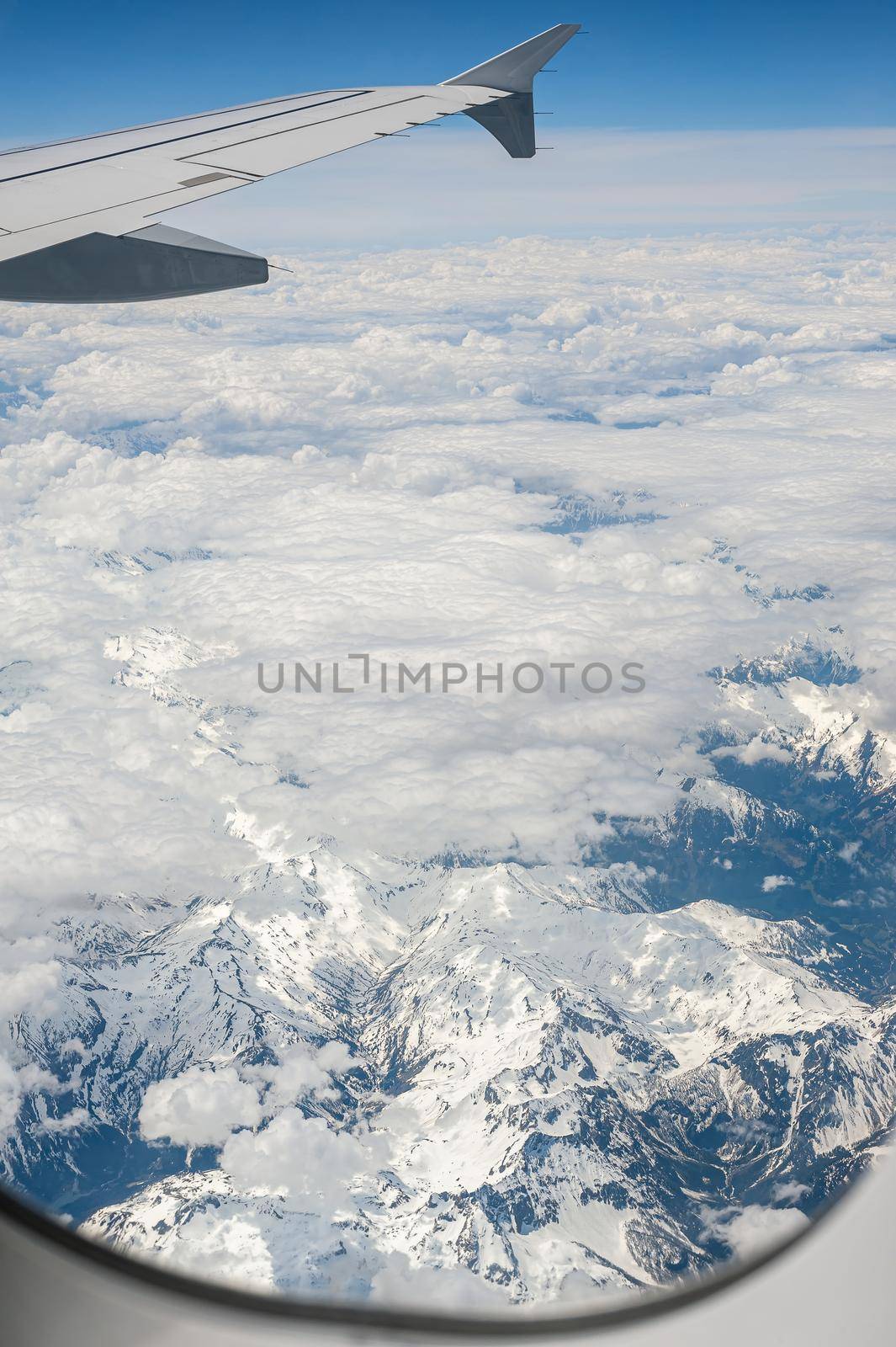 View from airborne airplane window at cloudy sky and Alps mountains with snow covered peaks from high altitude. by Qba