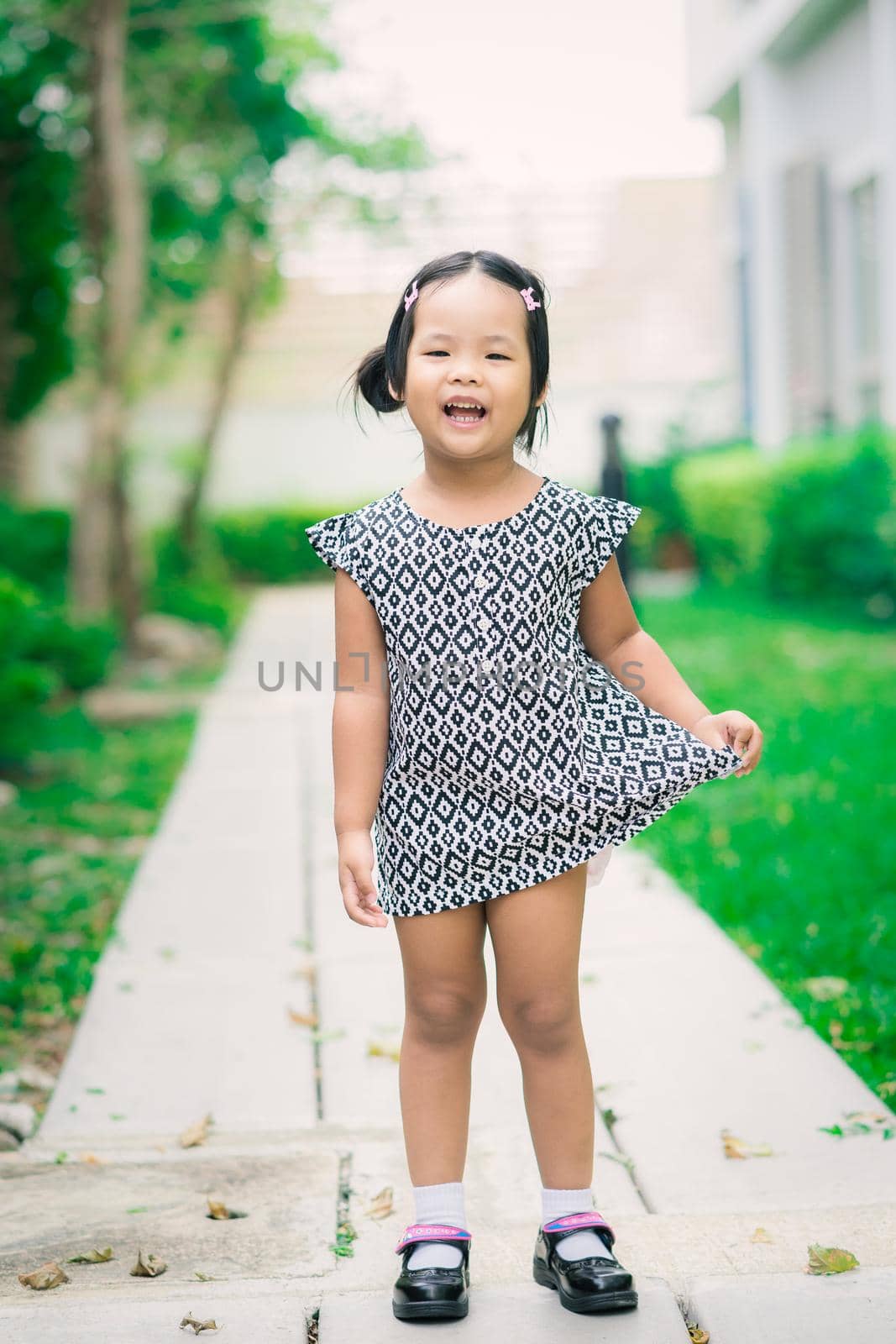 Portrait of happy asian little girl in dress standing on footpath in the park by domonite