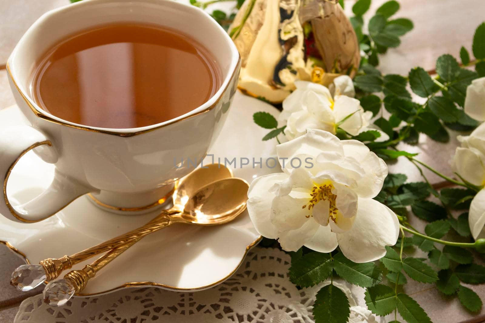 Cup of tea and branch of small white roses on rustic table, in white and golden colors