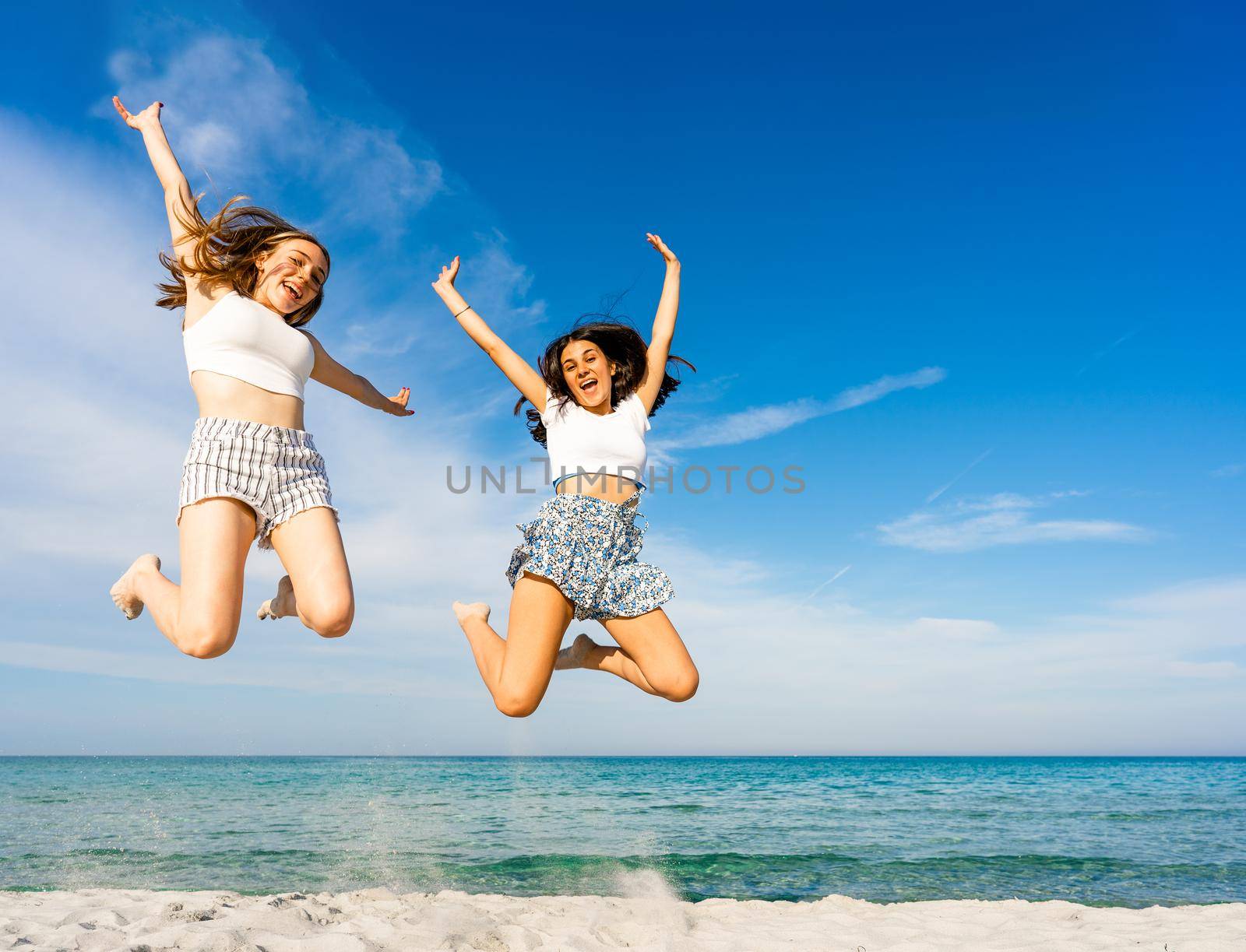 Two happy student girls jumping together looking at camera on tropical beach enjoying summer vacation in sea ocean resort. Happiness and success concept: find your talent to improve your life by robbyfontanesi