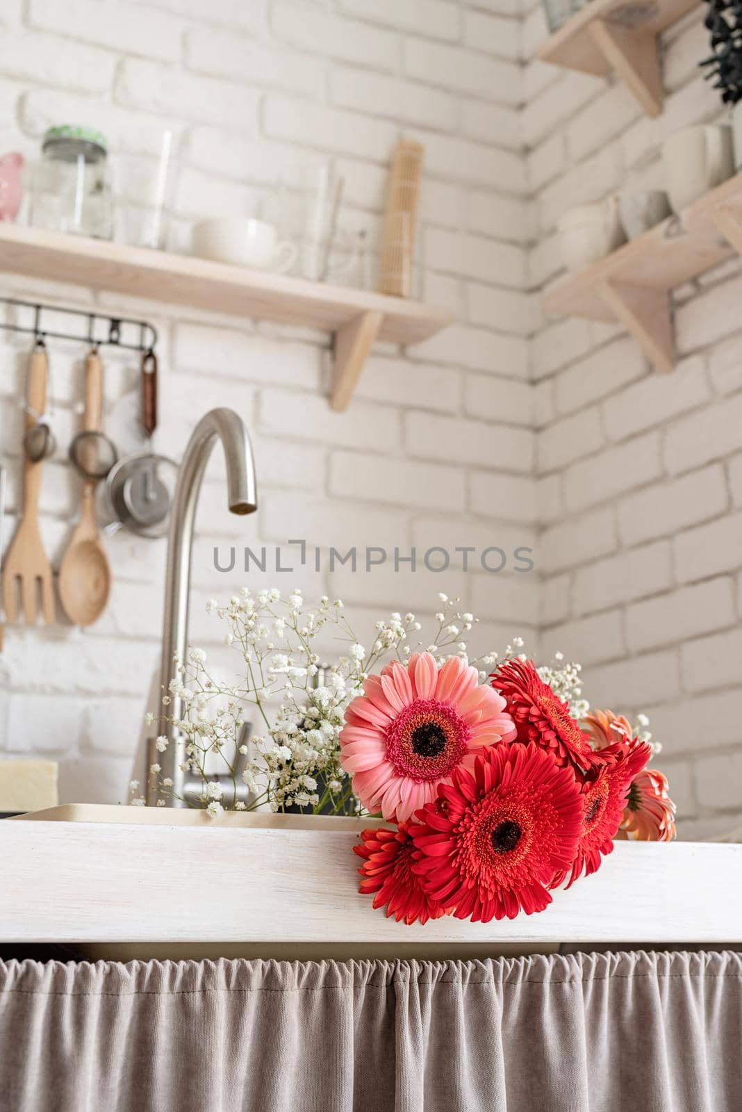 Rustic kitchen interior with white brick wall and white wooden shelves. Red and pink gerbera daisies in a kitchen sink