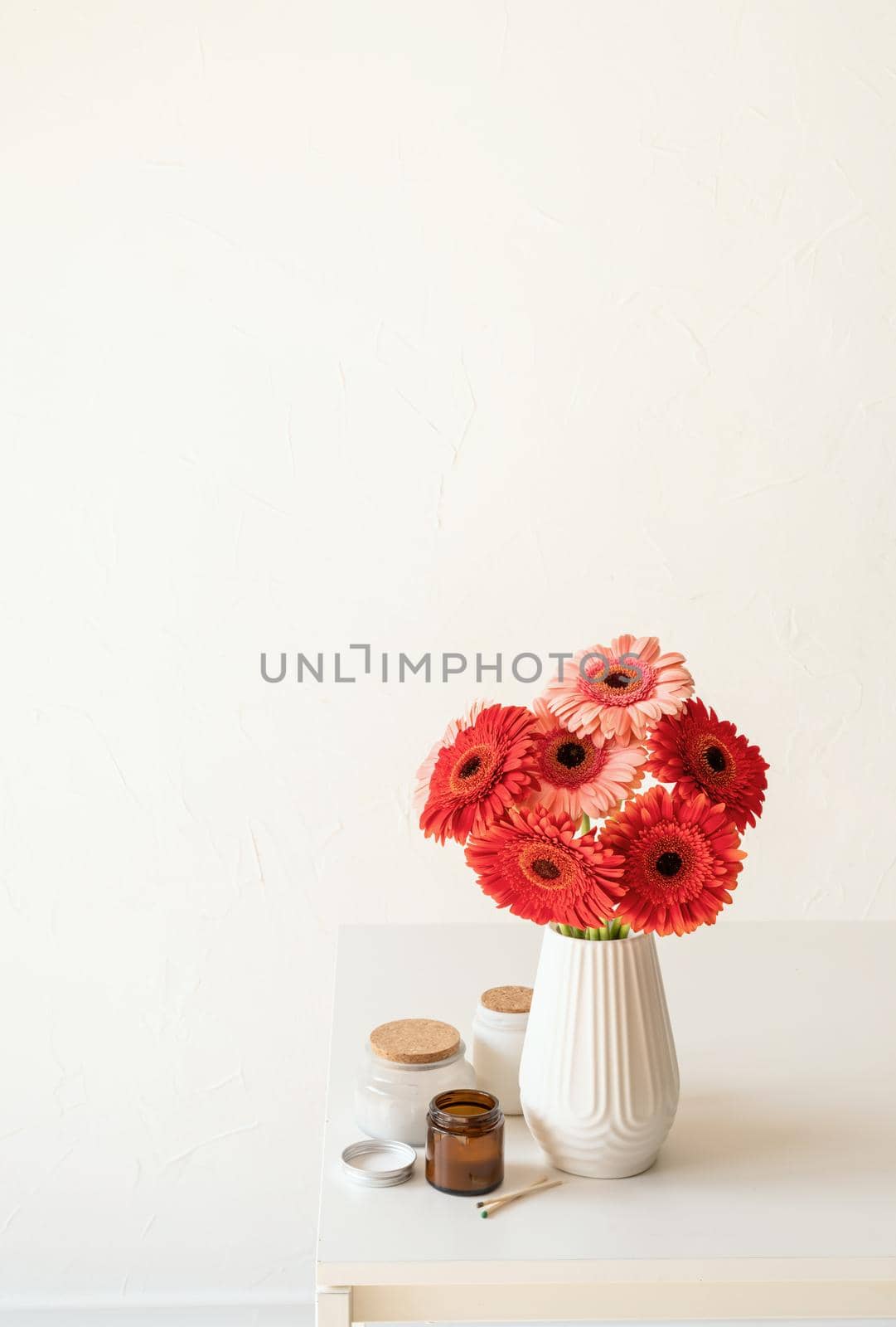Red and pink gerbera daisies in white vase on kitchen table with candles and cup, minimal style
