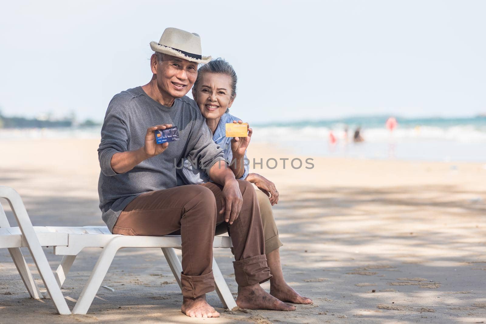 Happy senior couple smile sitting chair beach relaxing outdoors hold and showing credit card payment money buyer to recommending at the beach, plan life insurance at retirement couple concept