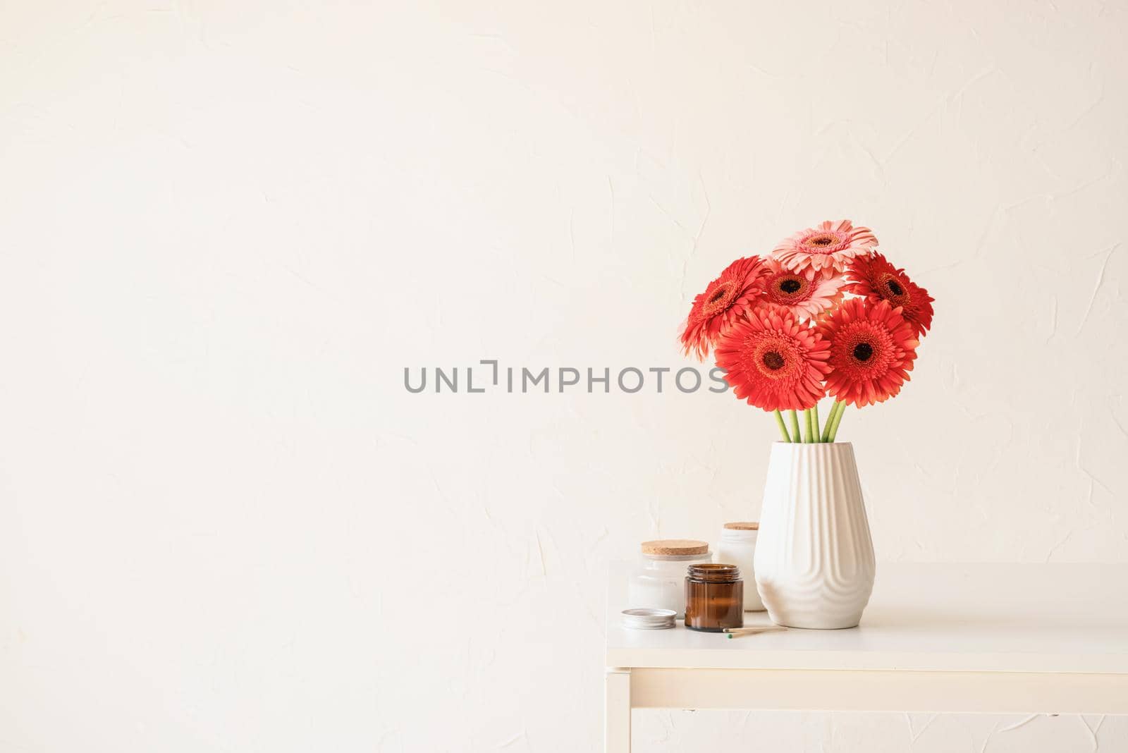 Red and pink gerbera daisies in white vase on kitchen table with candles, minimal style, copy space