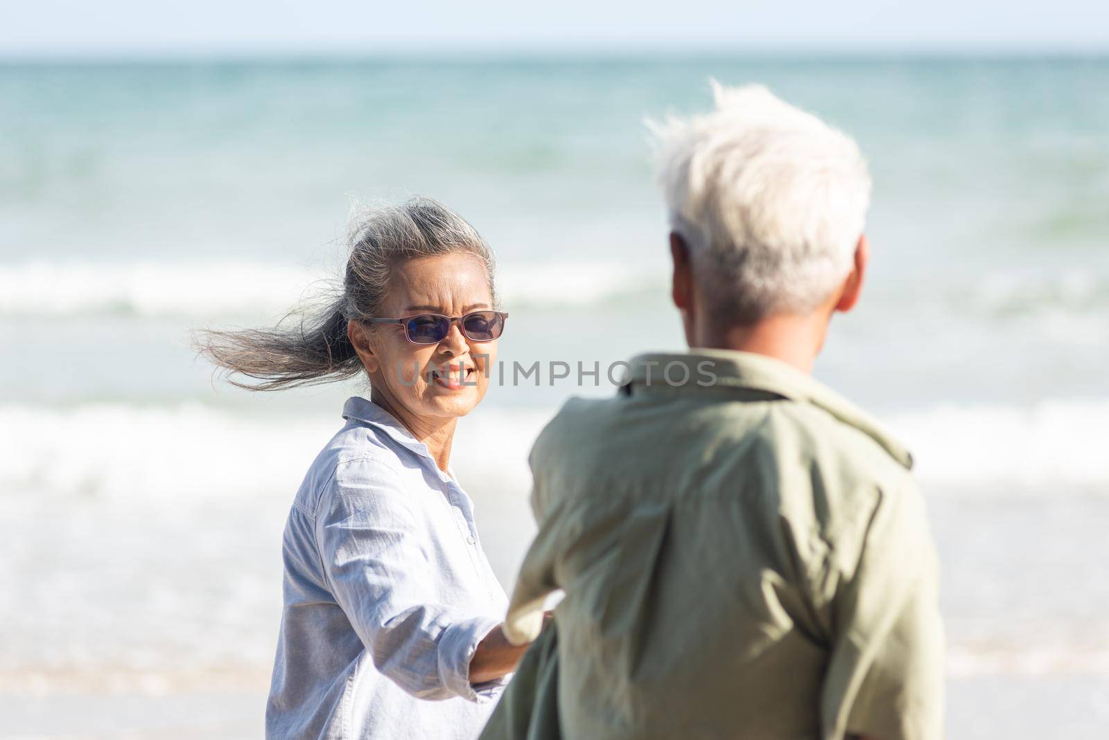 Happy Asian senior man and woman couple holding hands walking to the beach sunny with bright blue sky, Romantic elderly enjoy Travel summer vacation, plan life insurance at retirement couple concept