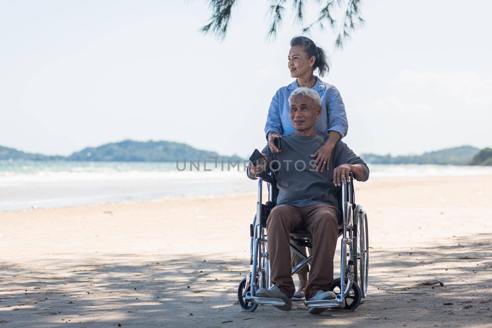 senior man on wheelchair and elderly woman couple smiling on sand of the beach by Sorapop