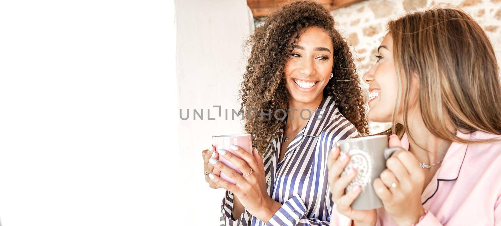 Multiracial women couple smiling looking each other holding a teacup in pajama just waken up. New normal gay families relationship and habits daily life scenes. Afro-American brunette curly woman by robbyfontanesi