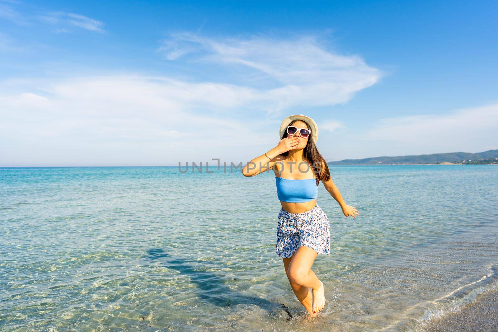 Young beautiful woman making kiss gesture with hand on mouth looking at camera walking in the seawater wearing white hat and sunglasses. Spend your time in travel for a happy better life experience