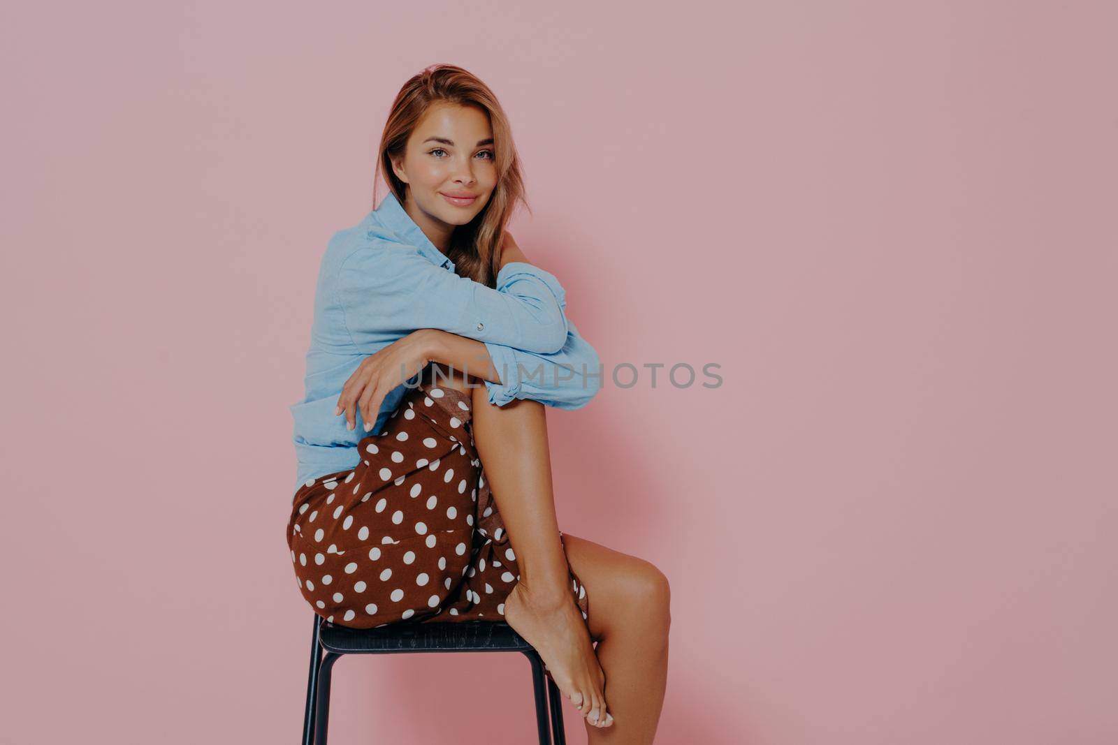 Young female model sitting sideways on black modern chair with one leg raised up. Beautiful sensual brunette female in casual shirt and polka dot skirt, isolated over pink studio background