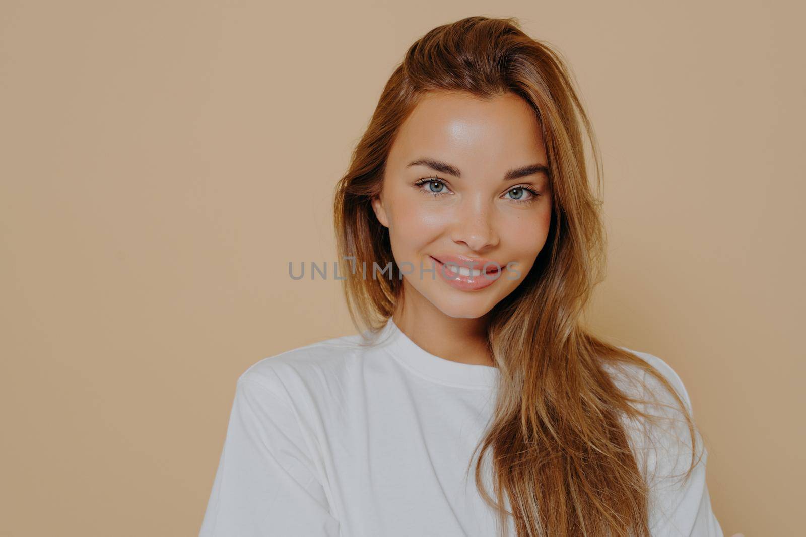 Portrait of beautiful female with long light brown straight hair gently smiling on camera wearing casual white tshirt while poses against pastel beige wall. Women beauty and positive emotions concept