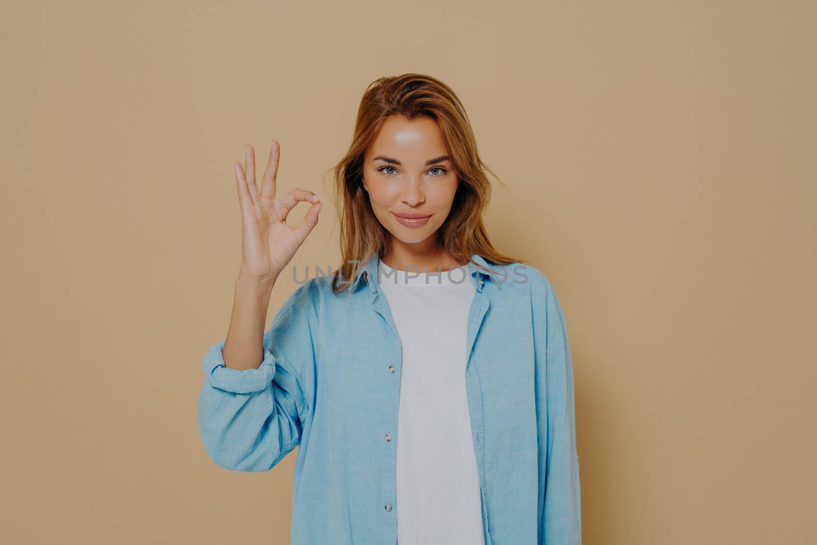 Charming young lady expressing approval or acceptance with okay gesture, confidently looking at camera and posing on beige background, female demonstrating agreement