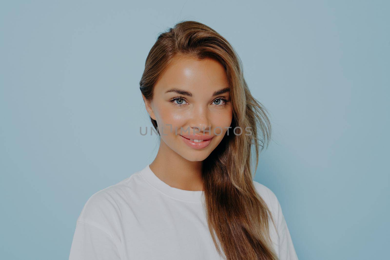 Close up portrait of smiling stylish young beautiful woman with long straight light brown hair in white t-shirt with natural light make up, isolated over blue studio background with copy space