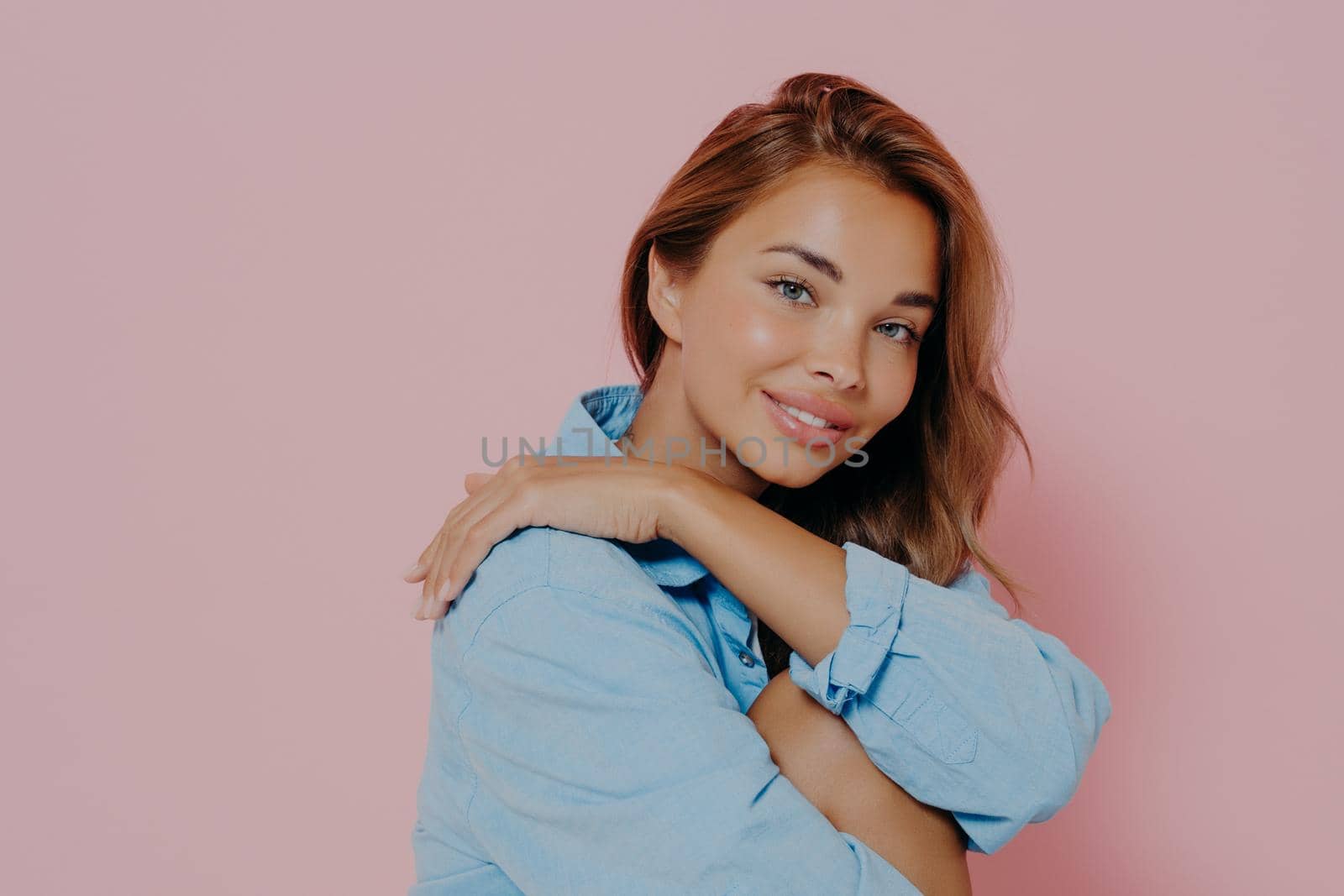 Charming young lady wearing stylish blue shirt with beautiful toothy smile, healthy hair and clean skin, feeling relaxed and comfortable while posing isolated over pink background