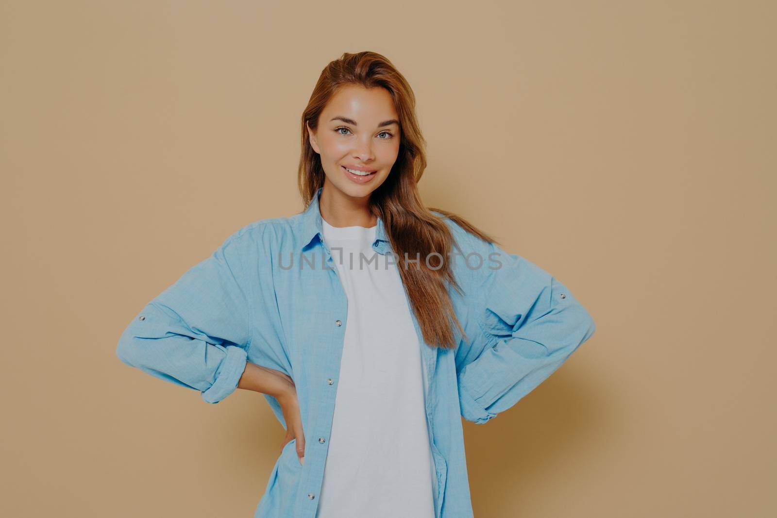 Self assured smiling lovely model keeps hands on waist, wears long blue shirt over white tshirt, looks directly at camera, expresses her right, isolated over beige background. Self confidence concept