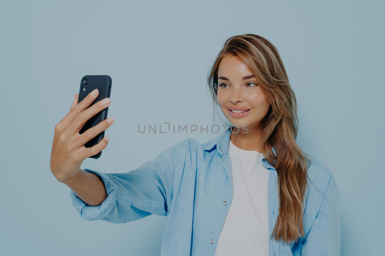 Time for selfie. Magnificent young woman in stylish casual clothes with long dyed hair taking photo of herself on modern cell phone, looking charming and flirty while posing on light blue background
