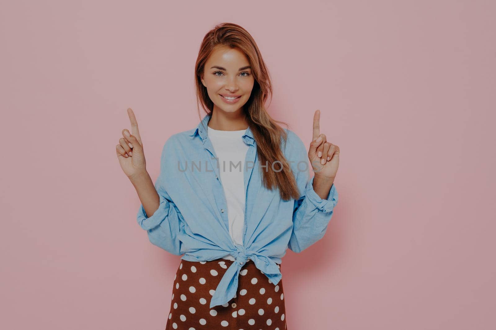 Young beautiful european woman with long stright hair wearing blue casual shirt and brown skirt with white polka dots, confidently pointing up with index fingers and smiling at camera