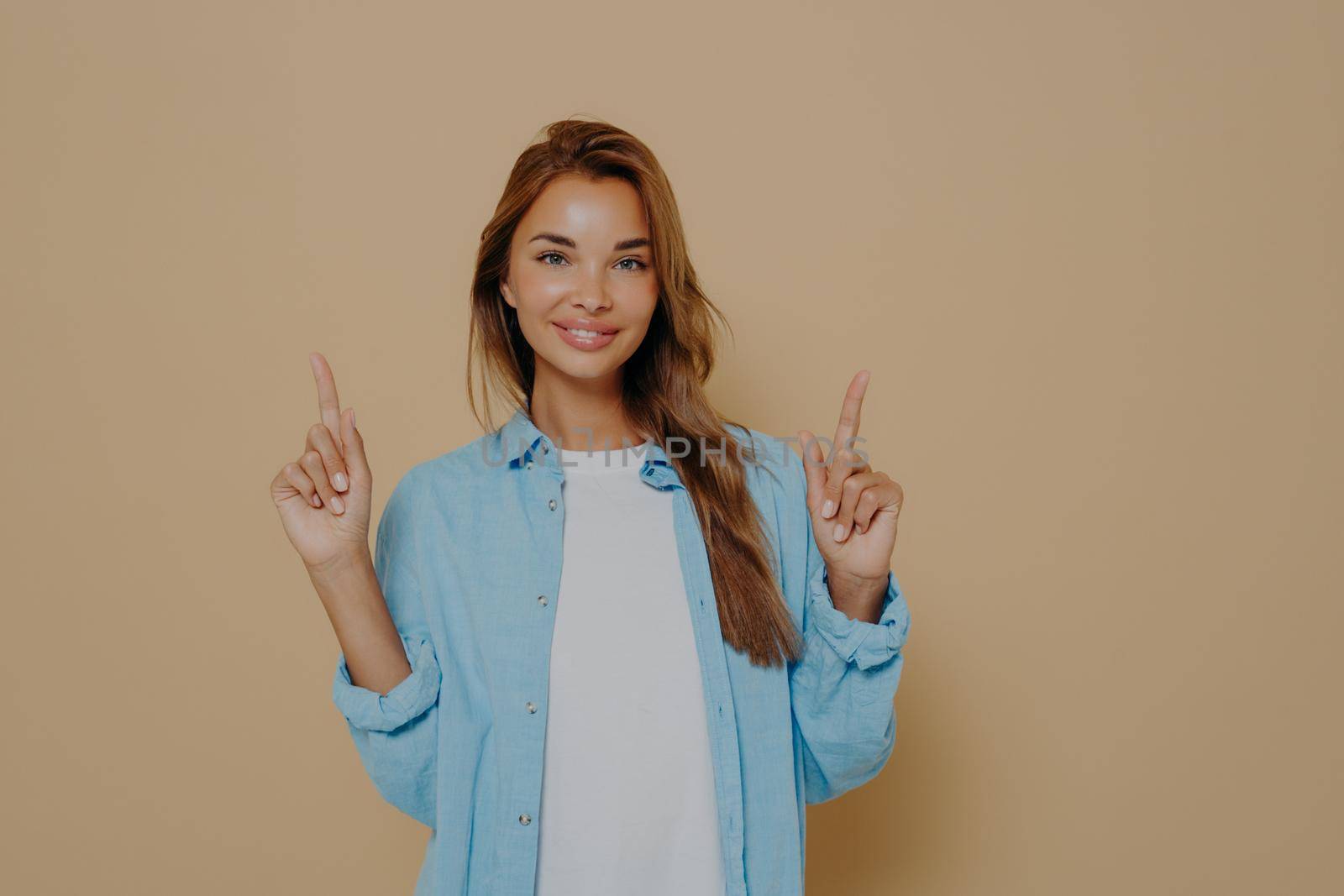 Advertising concept. Excited cheerful european female with long brunette hair wearing casual clothes and smiling happily, pointing index fingers up against beige background, attracting customers