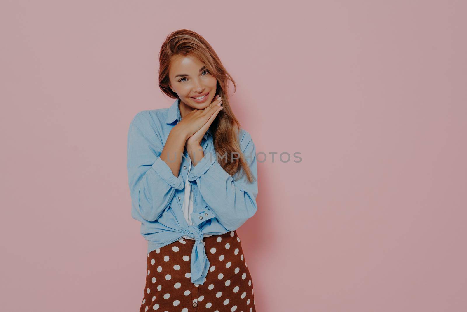 Attractive young woman in stylish outfit with beautiful sincere smile feeling love and cuteness, being shy and innocently looking at camera while standing against pink background