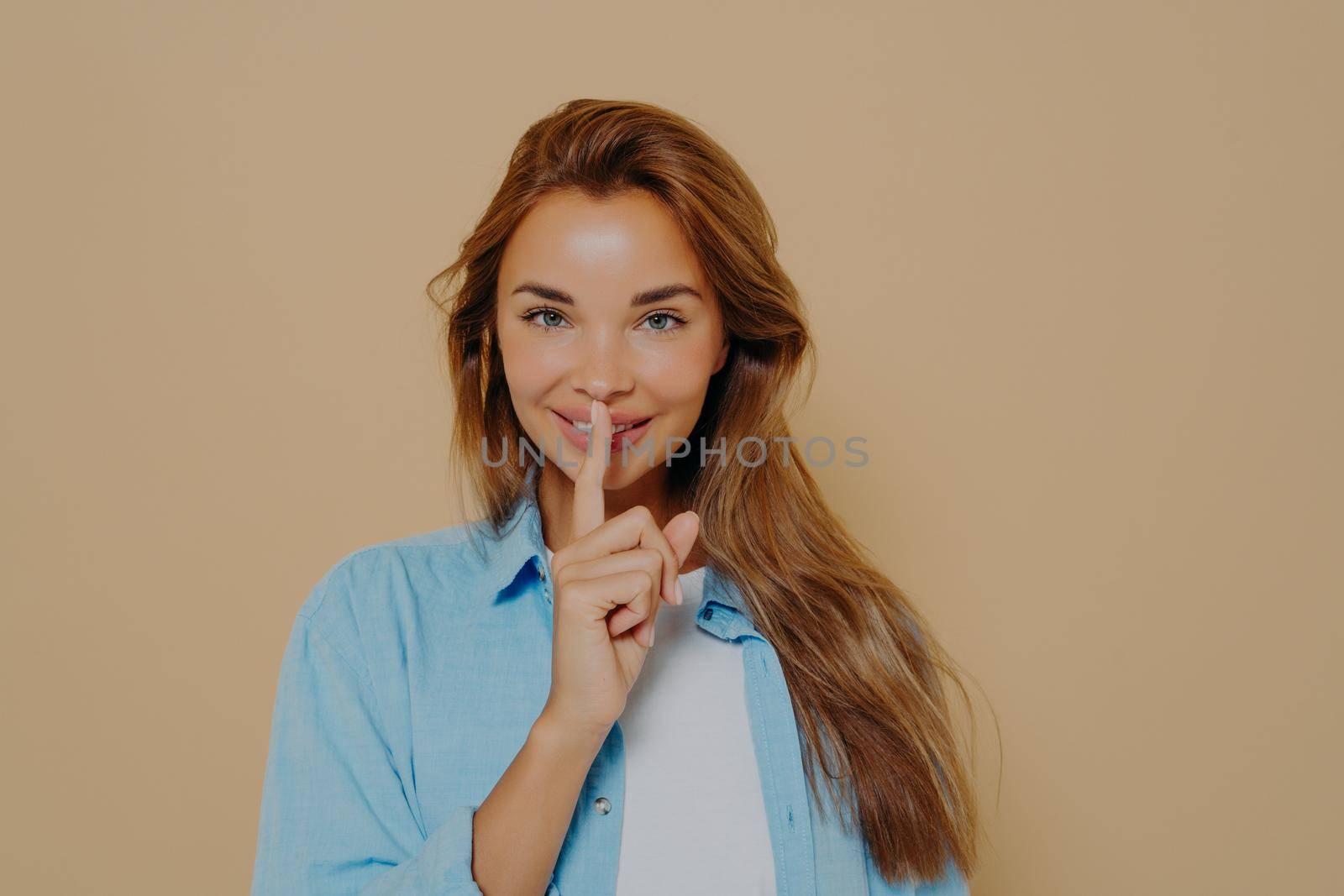 Portrait of joyful smiling female with long light brown hair holding index finger at her lips, saying 'shh', asking for silent or to keep her secret. Human face expressions, emotions and feelings