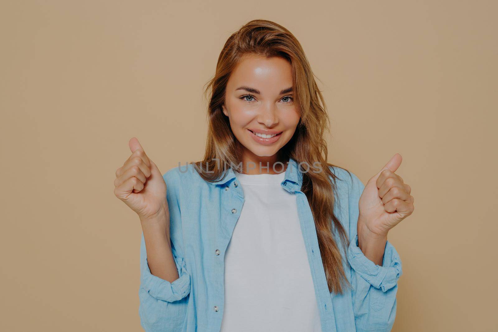 Overjoyed young european lady clenches fists with triumph, smiles with happiness, rejoices success, wears casual outfit cheers for favorite team, isolated on beige background