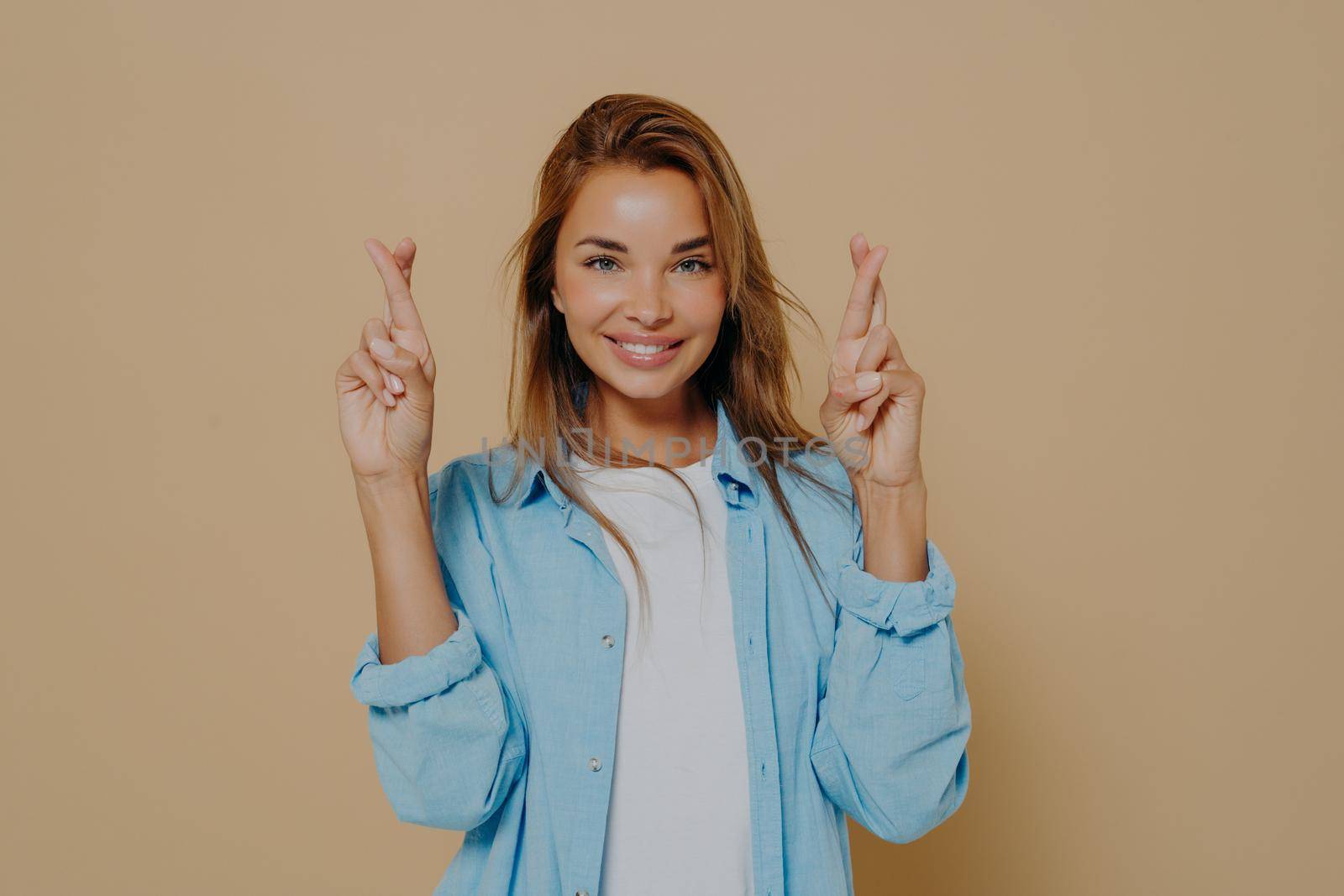 Positive superstitious young lady with beautiful smile holding her fingers crossed, making believer gesture, hoping for good luck and fulfillment of desire on beige background