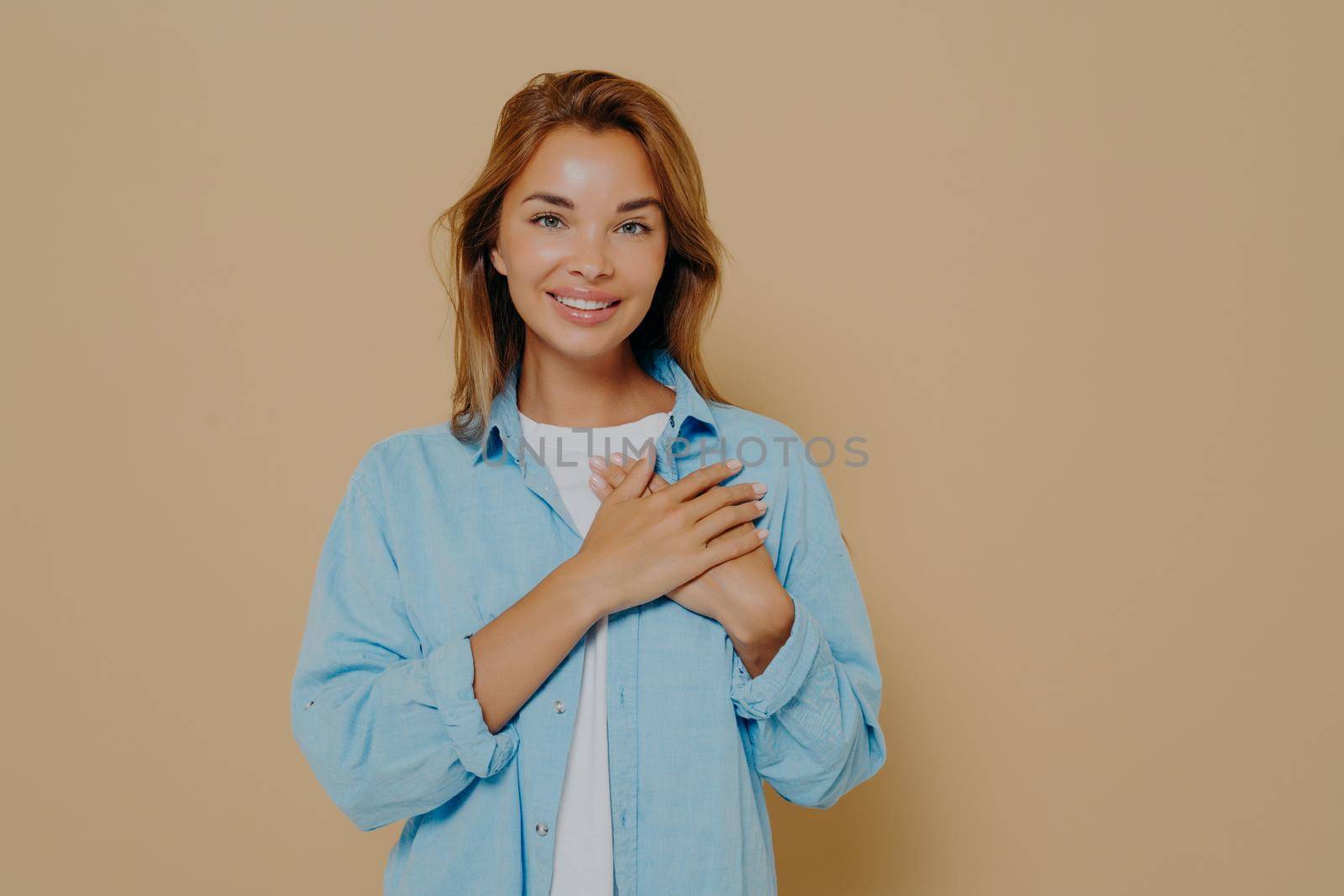 Delighted beautiful woman demonstrating her touched lovely mood by holding hands near heart, looking at camera with love and gratitude while posing in casual outfit on beige background