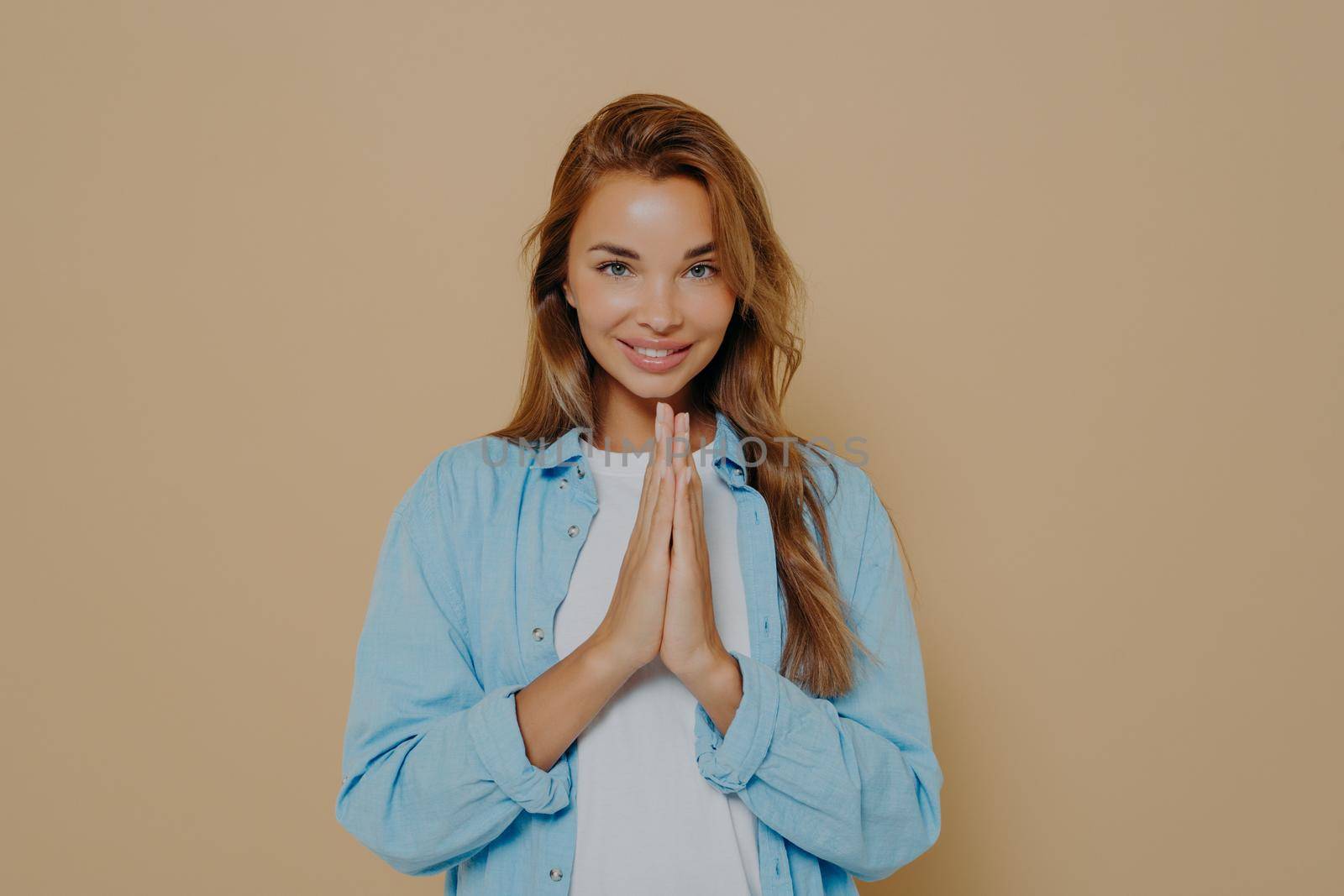 Pleased attractive woman putting her palms together in hope and prayer gesture and wishing for best, looking smiling at camera isolated on beige background. Body language concept