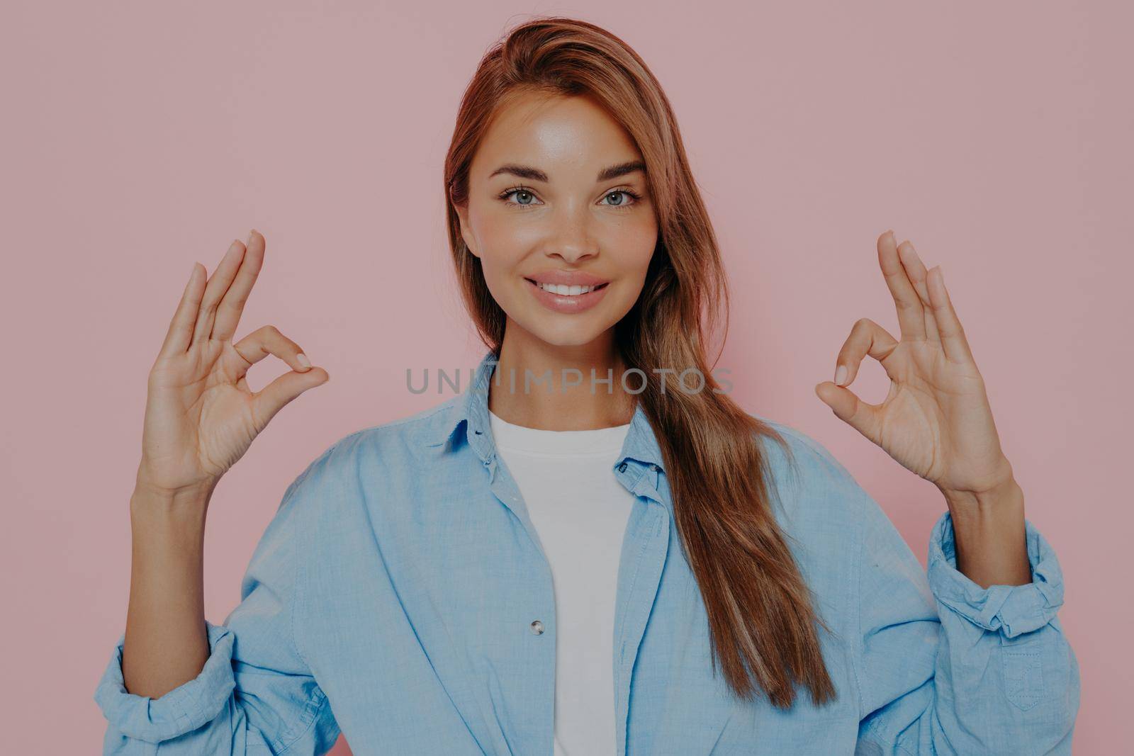 Happy young woman showing okay gesture with both hands and demonstrating aproval, looking at camera with cheerful smile, posing against pink background in stylish outfit. Body language concept