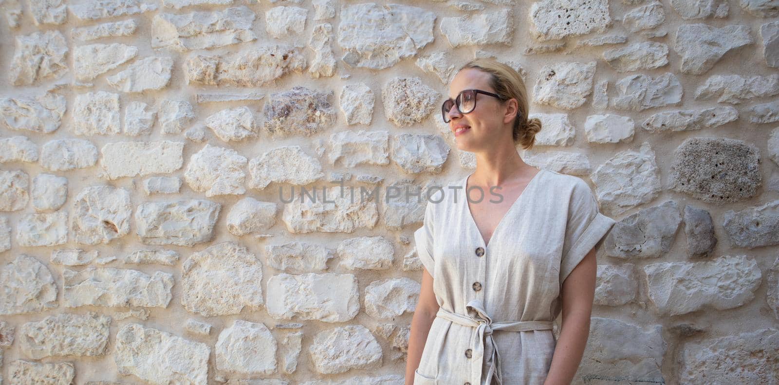 Portrait of beautiful cheerful blonde woman wearing one piece sundress and summer hat, standing in front of old medieval stone wall. Summer vacation portrait concept. Copy space.