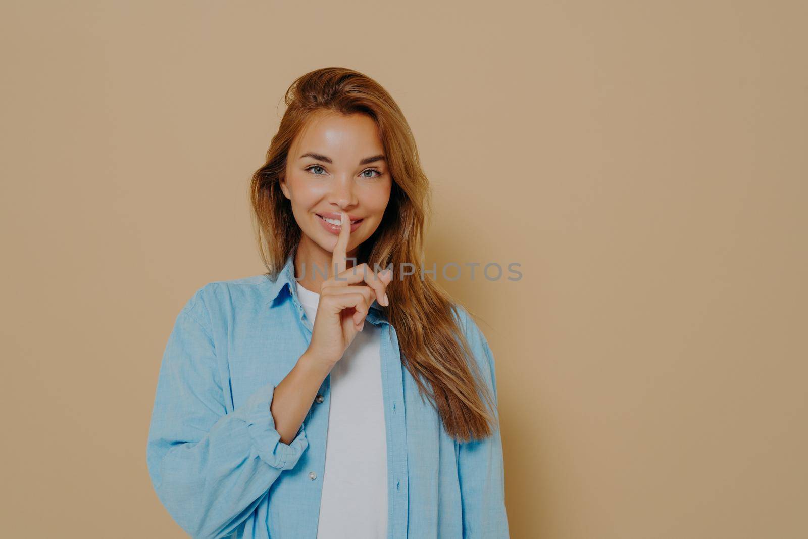 Positive attractive young woman wearing casual outfit, showing silence gesture, asking not to tell anyone secret information, being excited about what she knows while standing on beige background