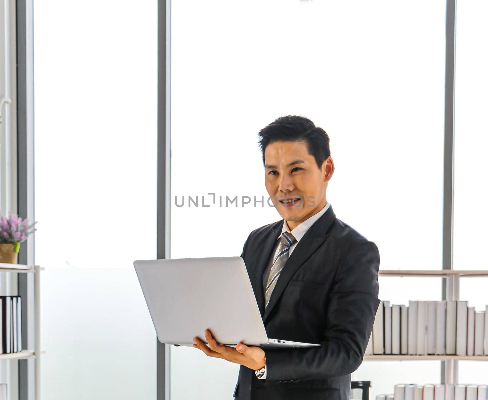 An Asian business man stands and holds a laptop computer to work at the office. Use apps for social media and online technology news for business strategies. Smile and be happy