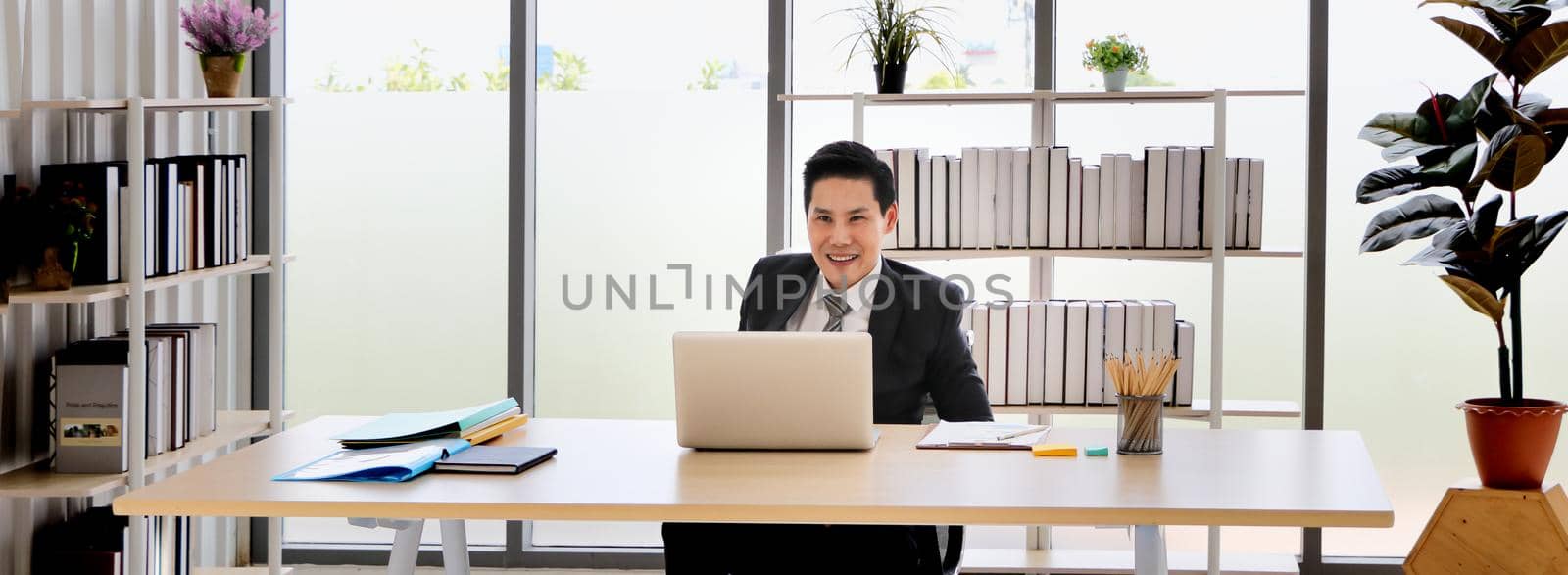 Portrait of a cheerful Asian businessman sitting at a desk looking at the PC screen. by atitaph